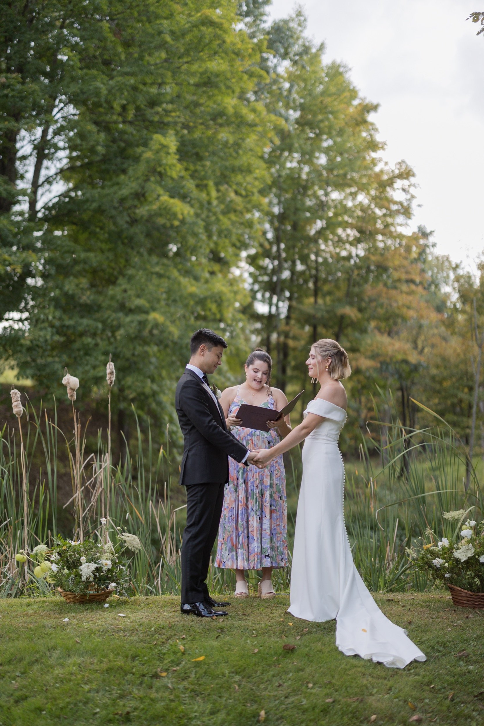 A bride and groom holding hands during their outdoor wedding ceremony, with a officiant standing behind them reading from a book, set in a lush green garden with trees and flowers.