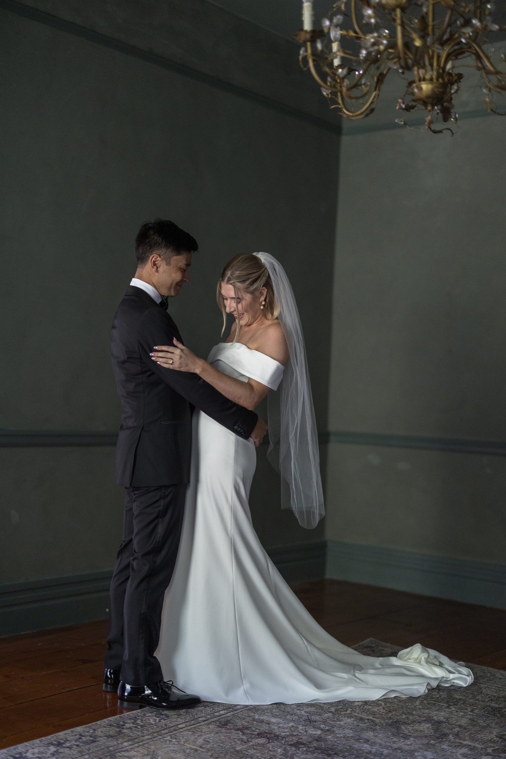 A bride and groom share a joyful moment during their wedding, standing on a wooden floor with a rug, in an elegant room with a chandelier overhead. The bride wears a white off-the-shoulder wedding gown and veil, while the groom is in a black tuxedo.