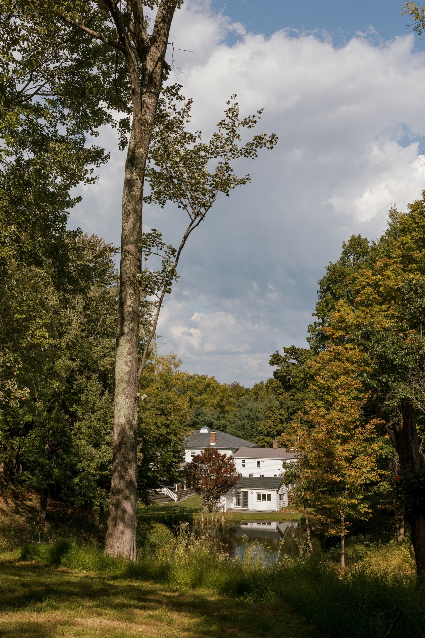 Scenic view of a white house surrounded by trees and a pond, with a partly cloudy sky overhead.