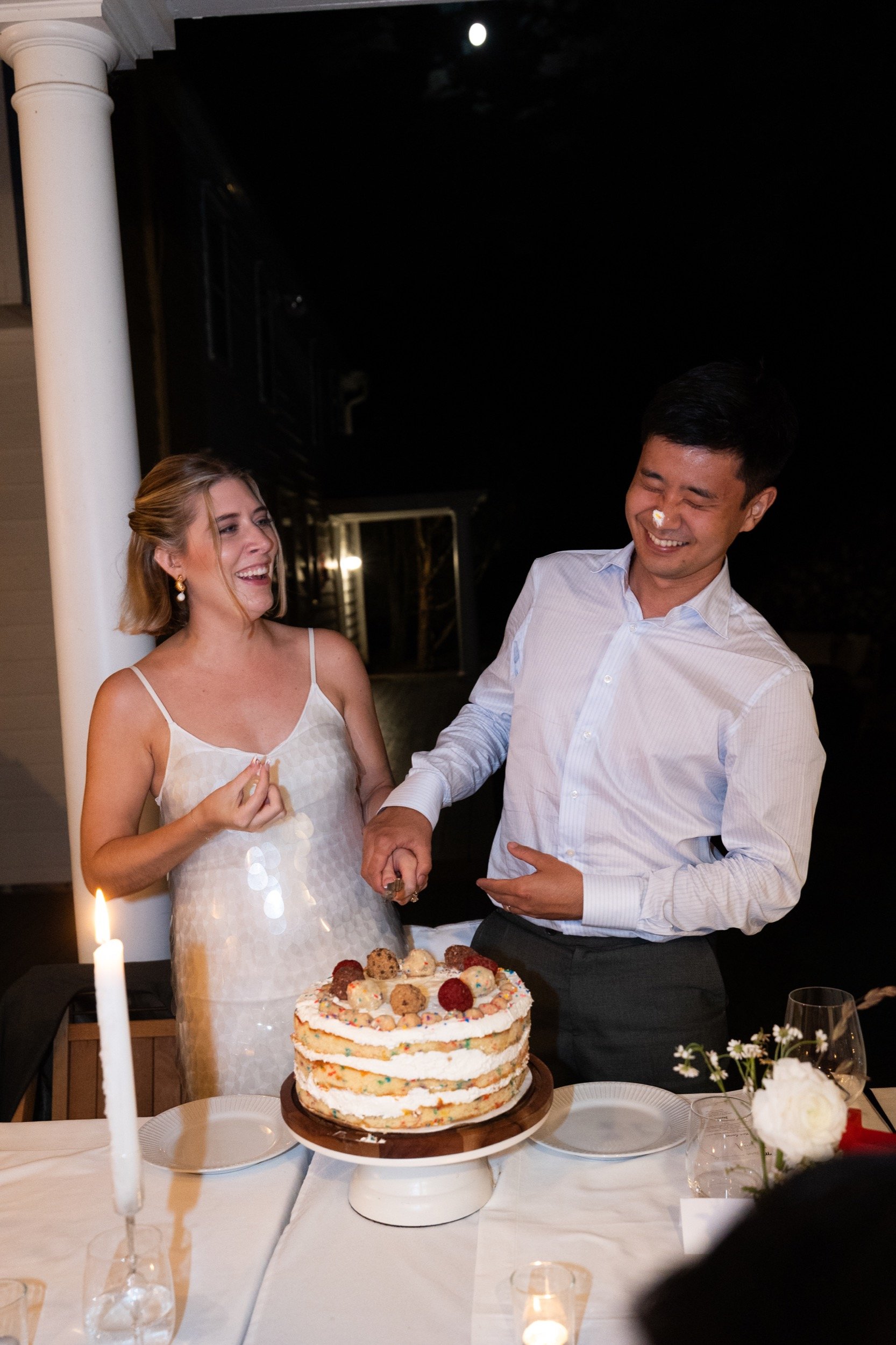 A woman and a man celebrating a birthday with a cake. The woman is smiling and holding hands with the man, who has a piece of cake on his nose. The cake is on a table with plates, candles, and flowers.