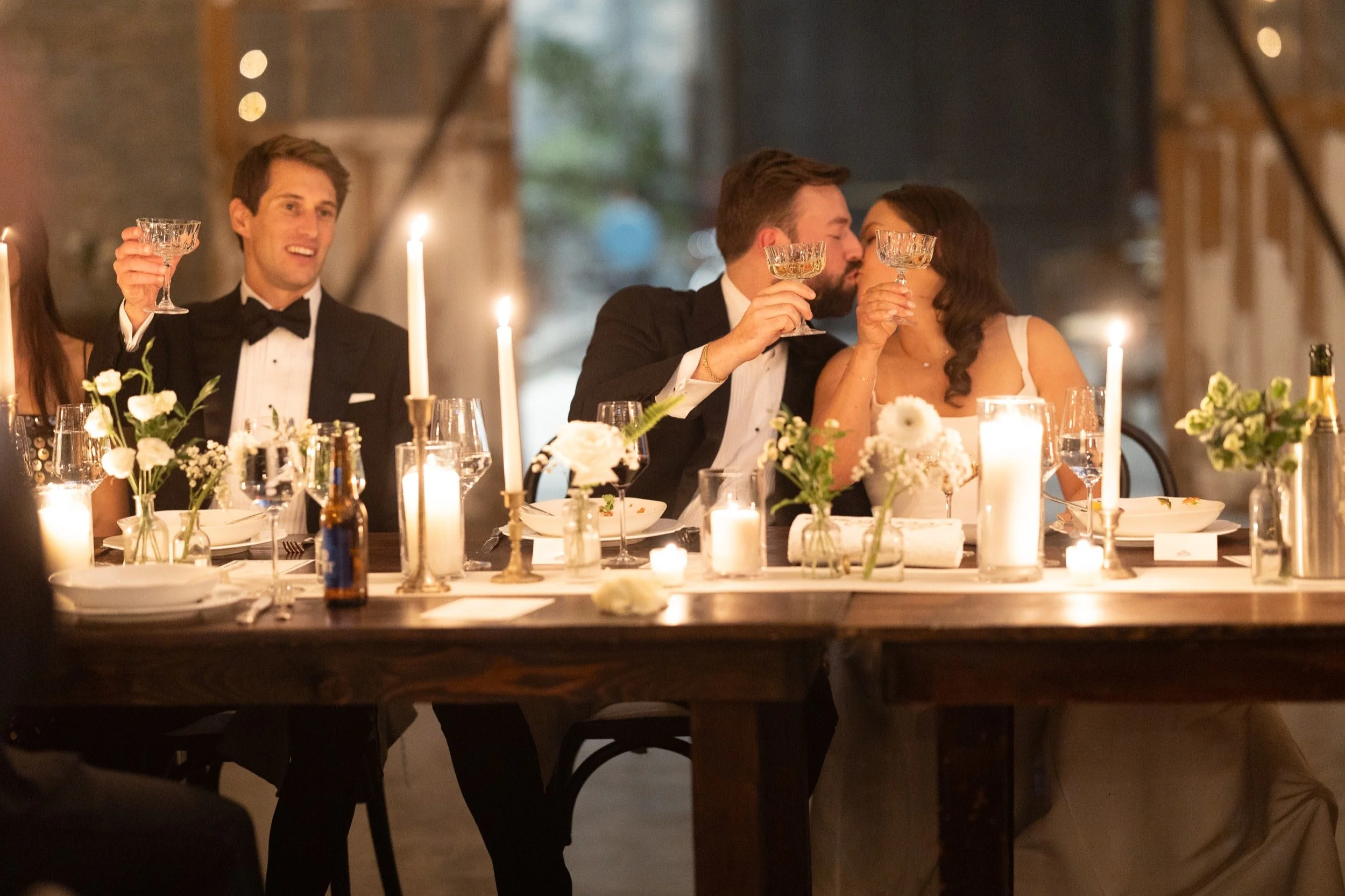 A candlelight Black tie wedding reception, where the bride and groom share a kiss after toasting during speeches.