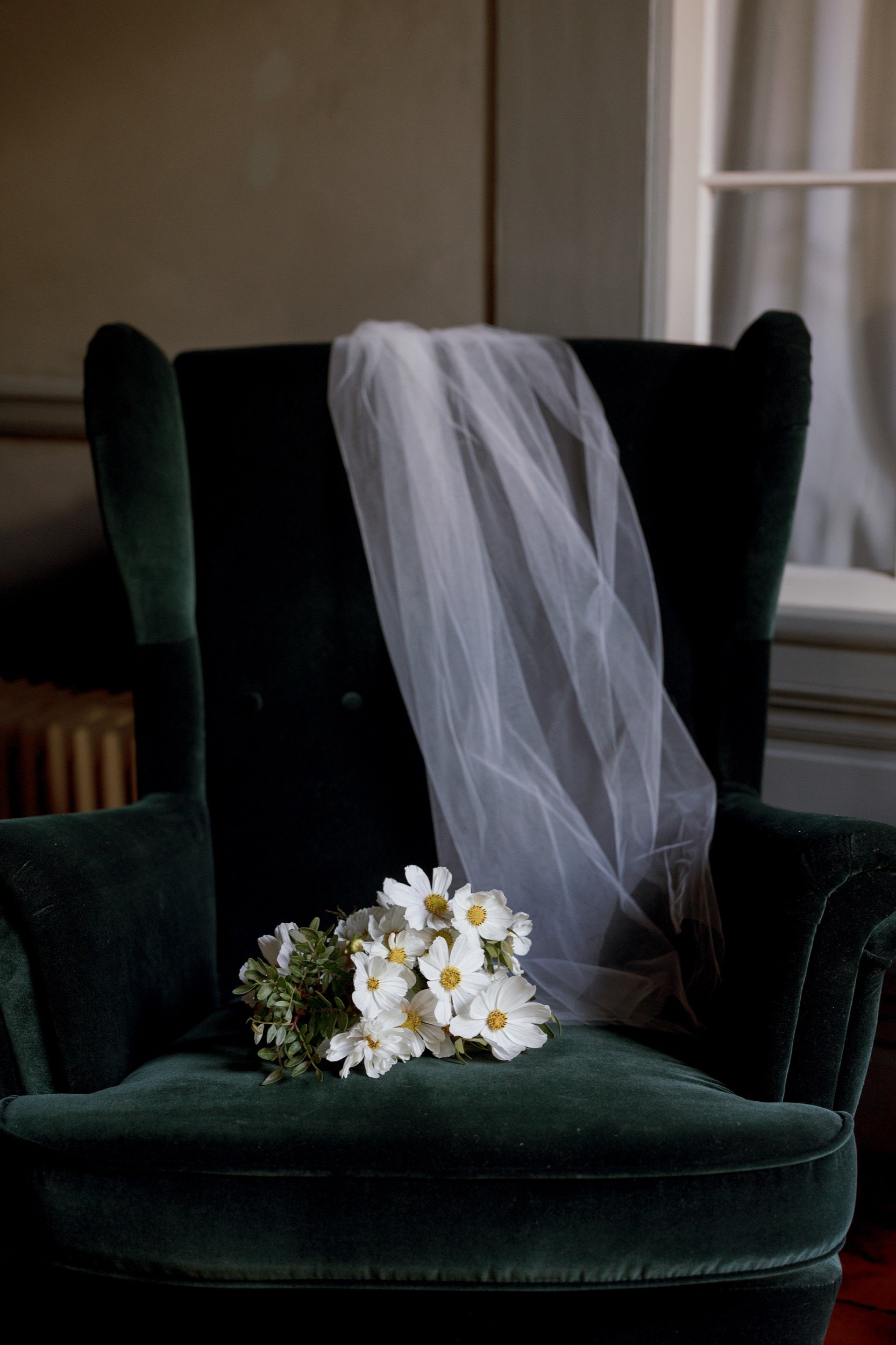 A dark green armchair with a white wedding veil draped over its back and a bouquet of white flowers resting on the seat.