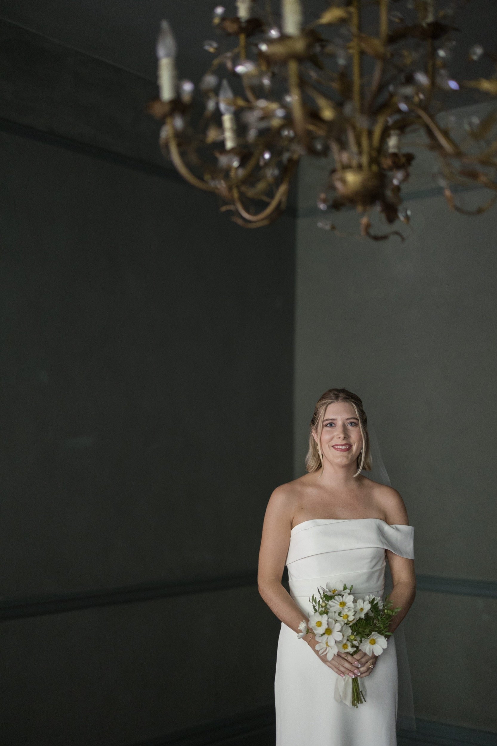 Bride in a white off-shoulder wedding dress holding a bouquet of white daisies, standing in a dimly lit room under a chandelier.