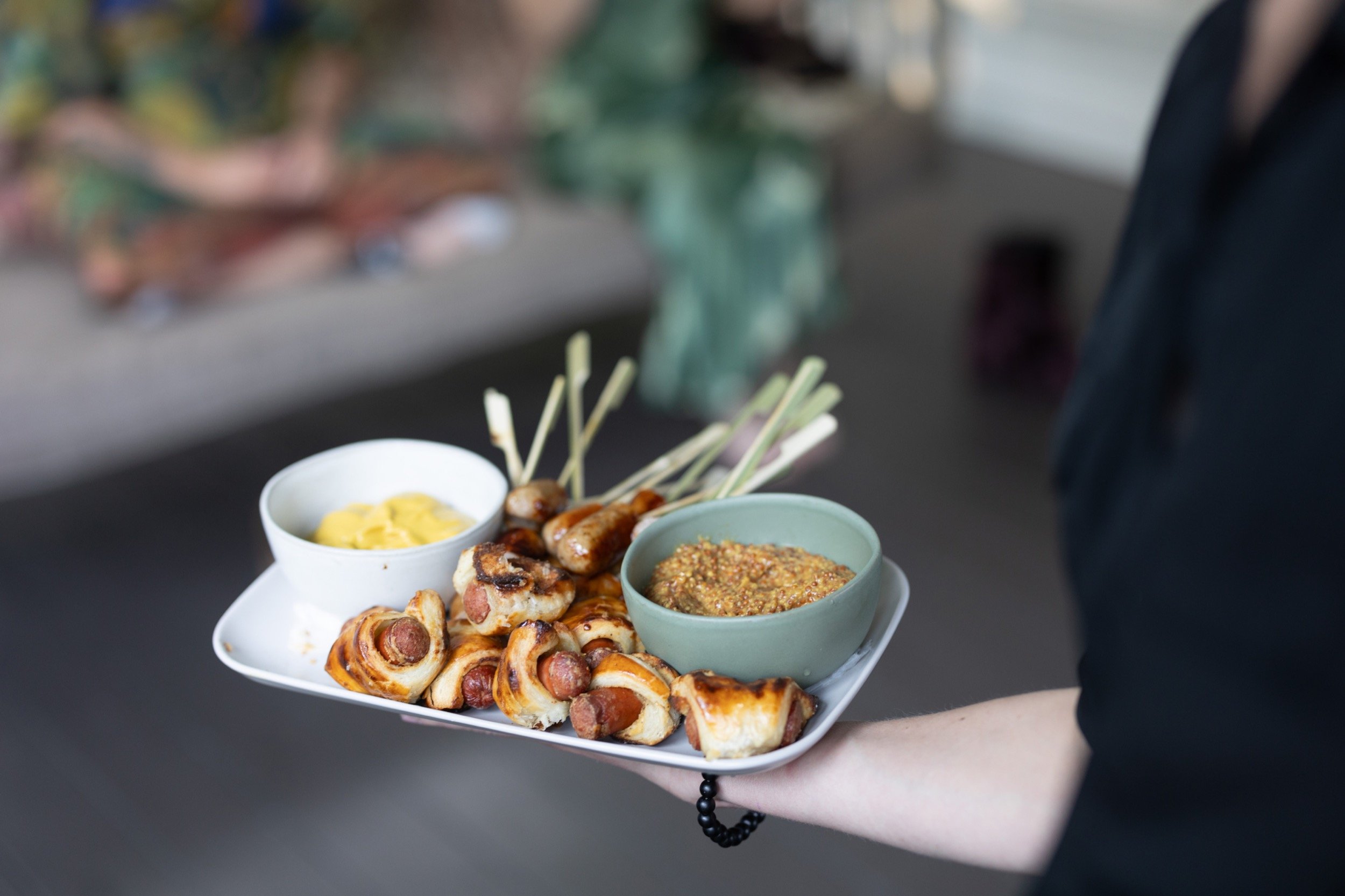 A person holding a white tray with grilled sausages, skewers of meat, mustard, and mustard dipping sauce with a blurred background.