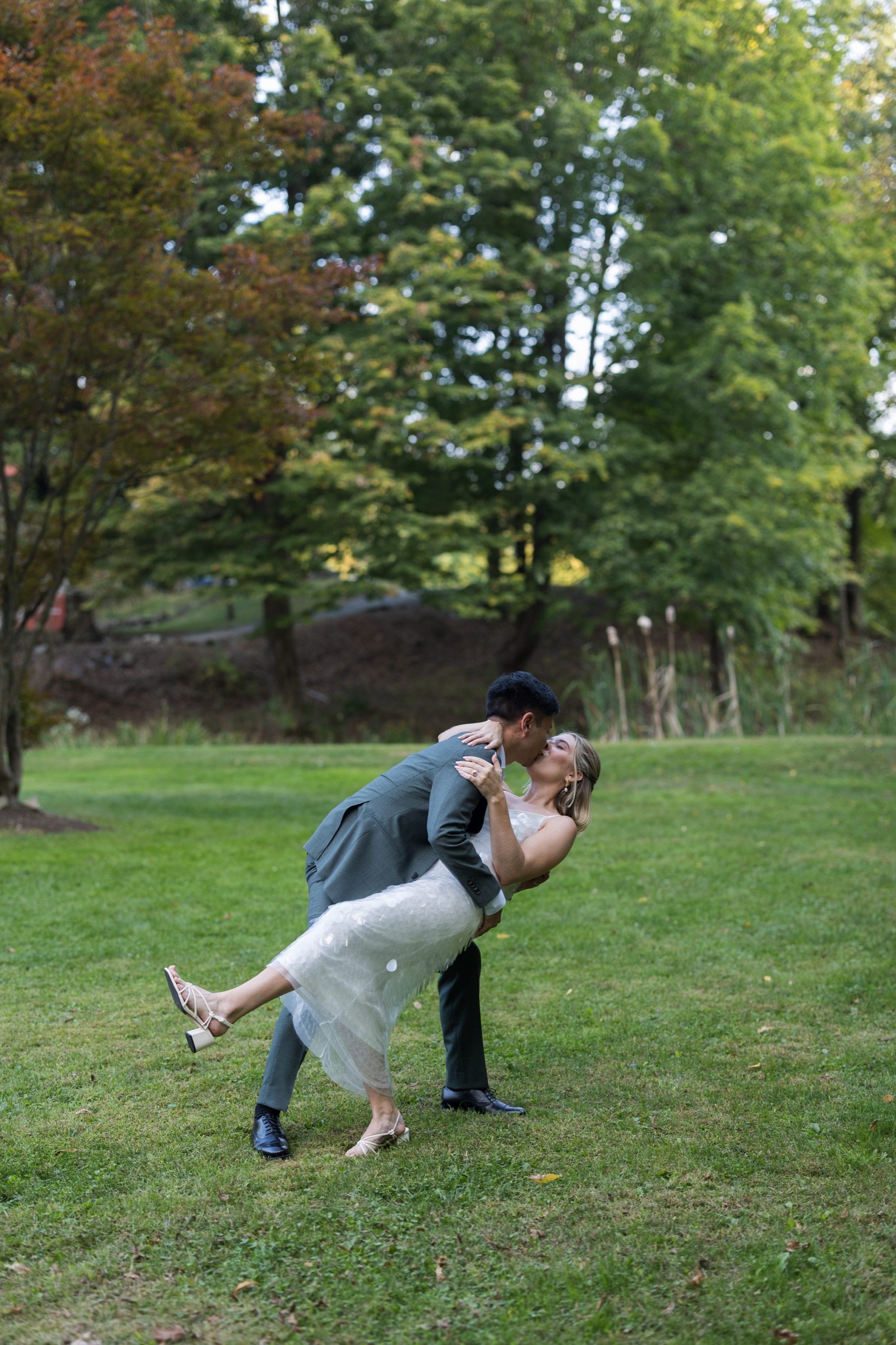 A man in a gray suit dips a woman in a white dress as they kiss on a grassy field with trees in the background.