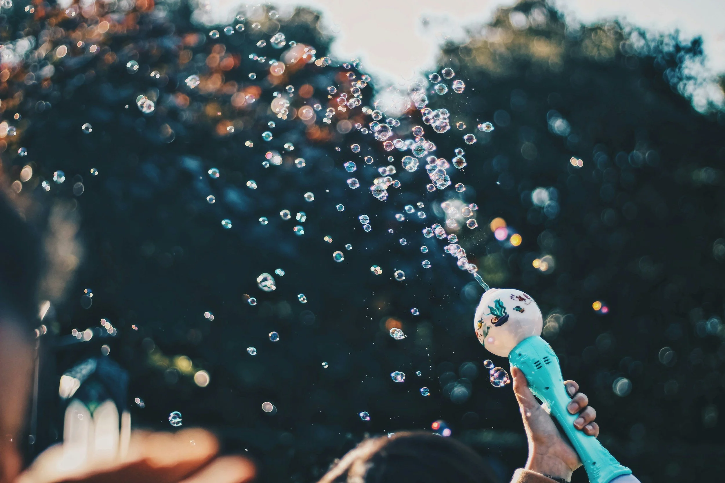 A person holding a bubble machine, creating bubbles outdoors with sunlight filtering through trees.