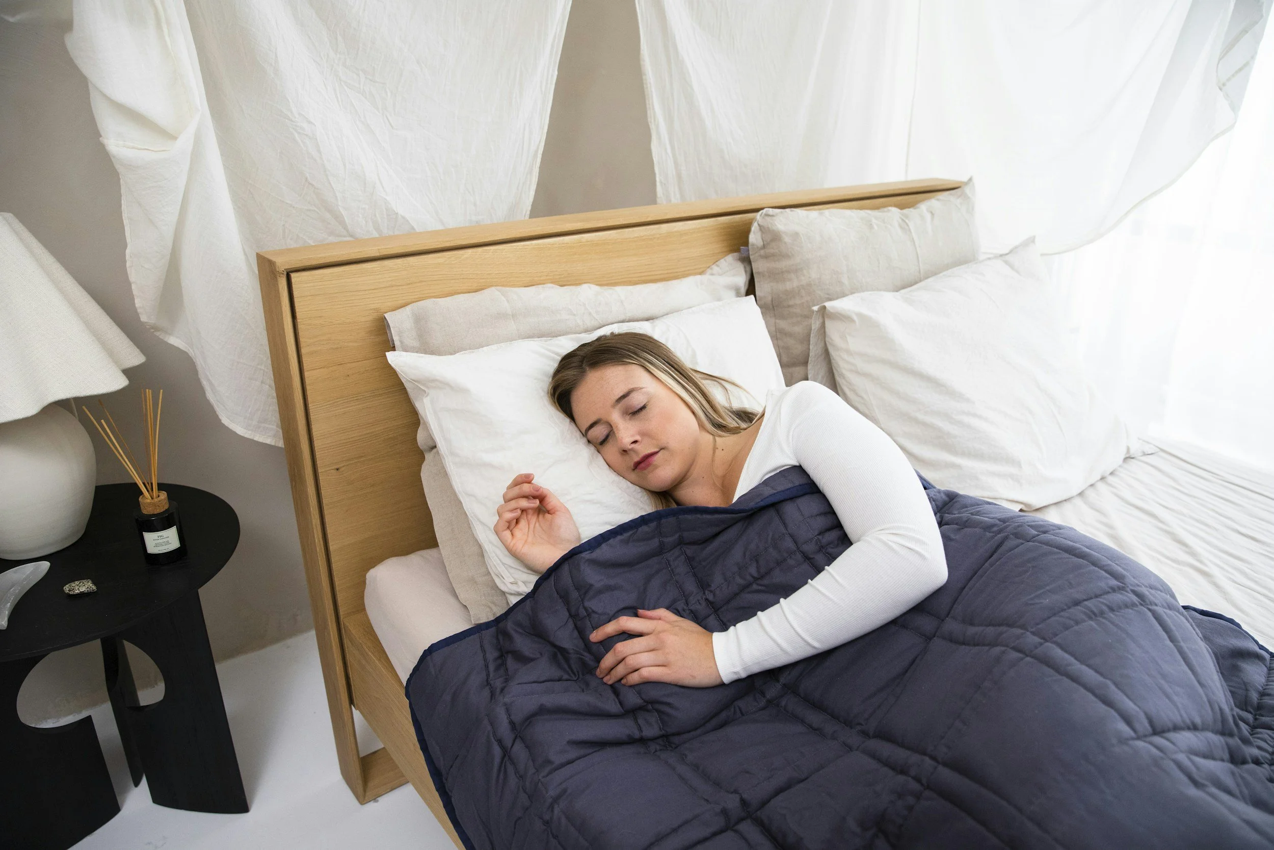 A woman sleeping in a bed with multiple pillows and a dark blue quilt, in a room with white curtains and a nightstand with a lamp and diffuser.
