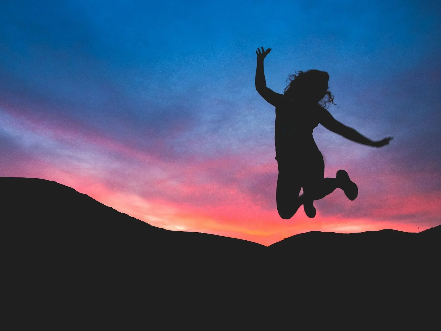 Silhouette of a person jumping in the air during sunset with colorful sky and mountain silhouettes in the background.