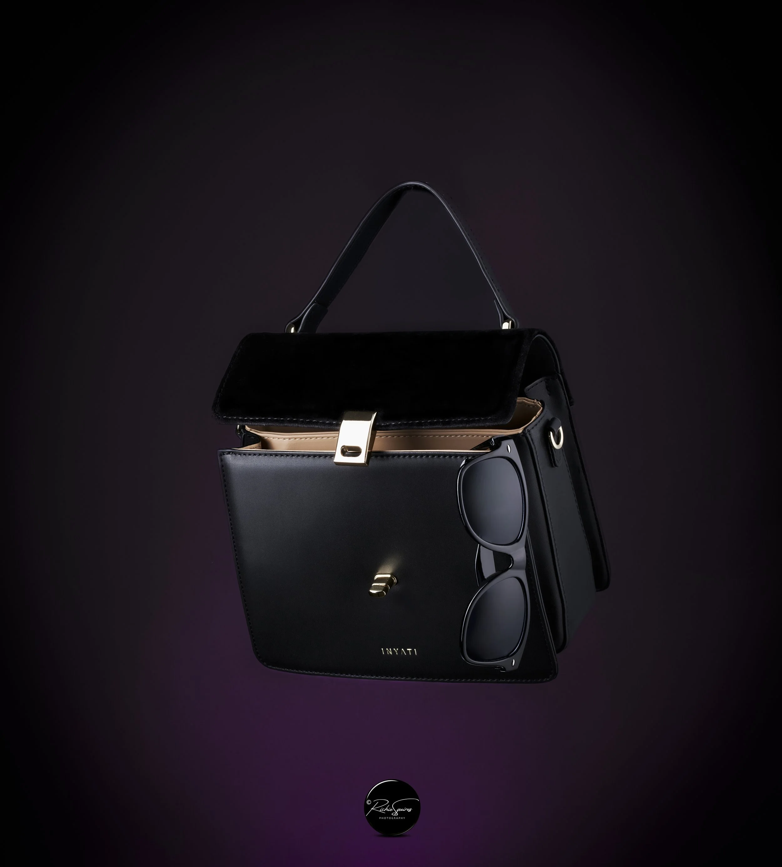 Black leather handbag with gold clasp and sunglasses positioned on front, set against a dark background.