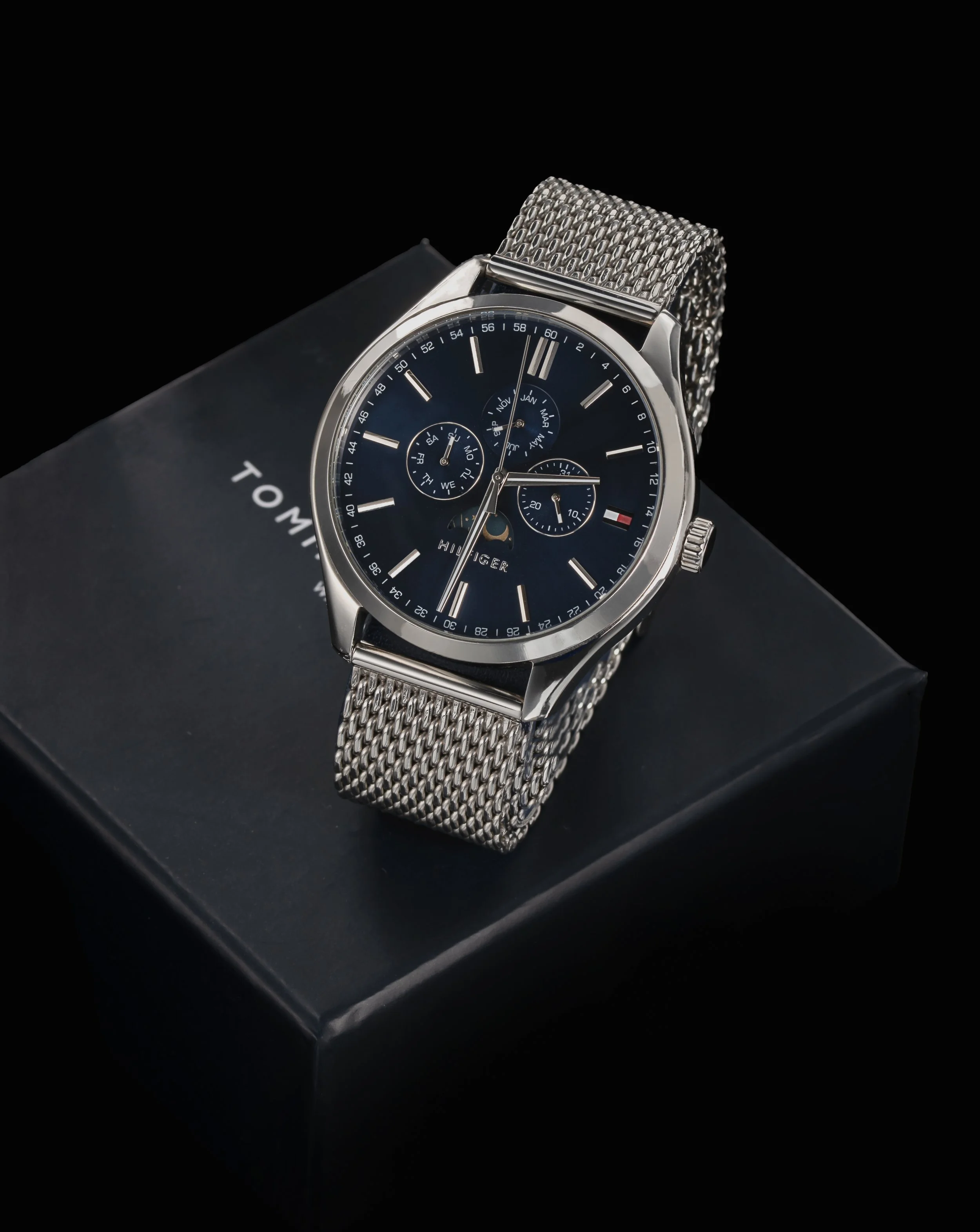 Luxury wristwatch with a blue dial, silver mesh band, and chronograph features displayed on a black box.