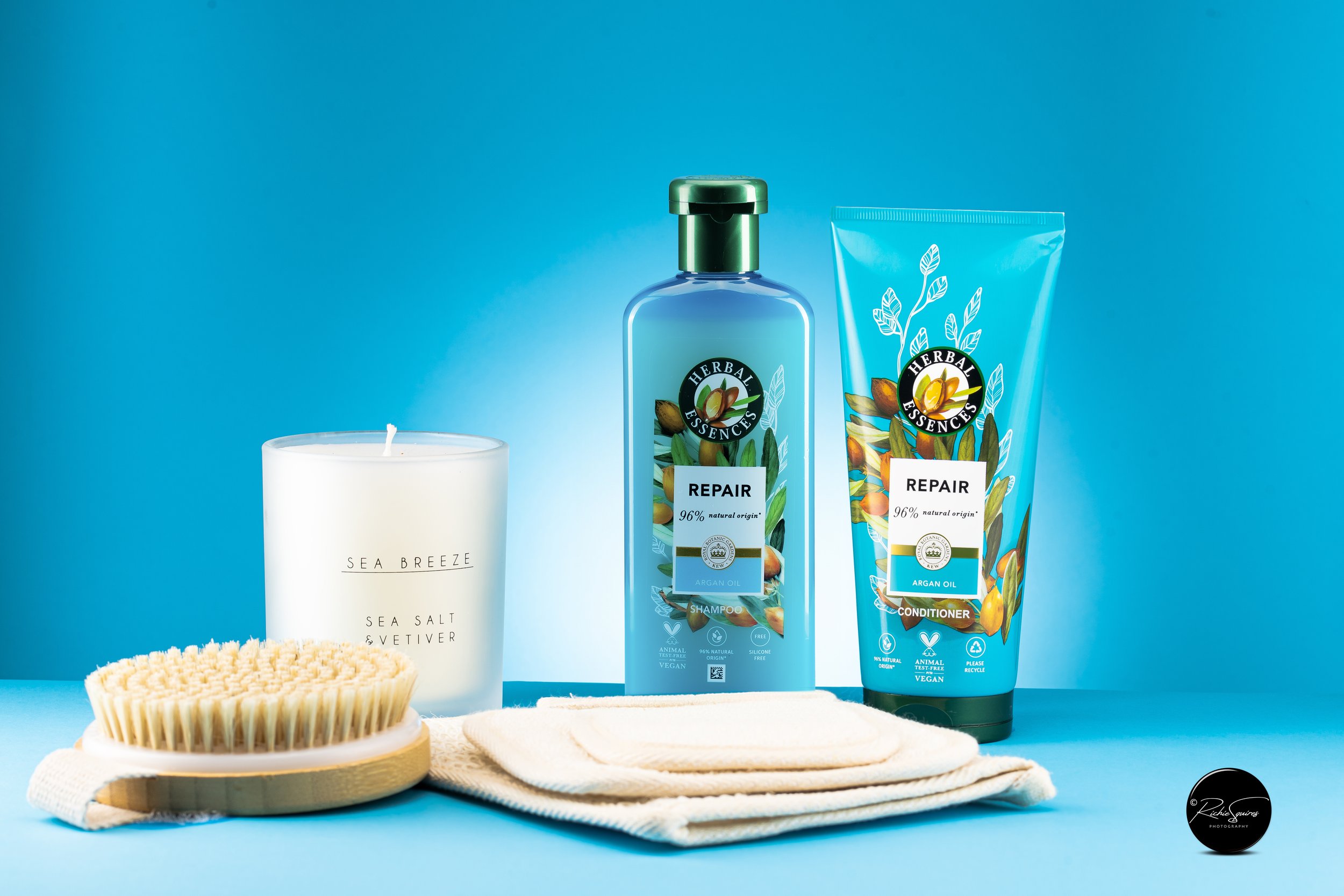 Bath and body products including shampoo, conditioner, candle, and brush against a blue background.