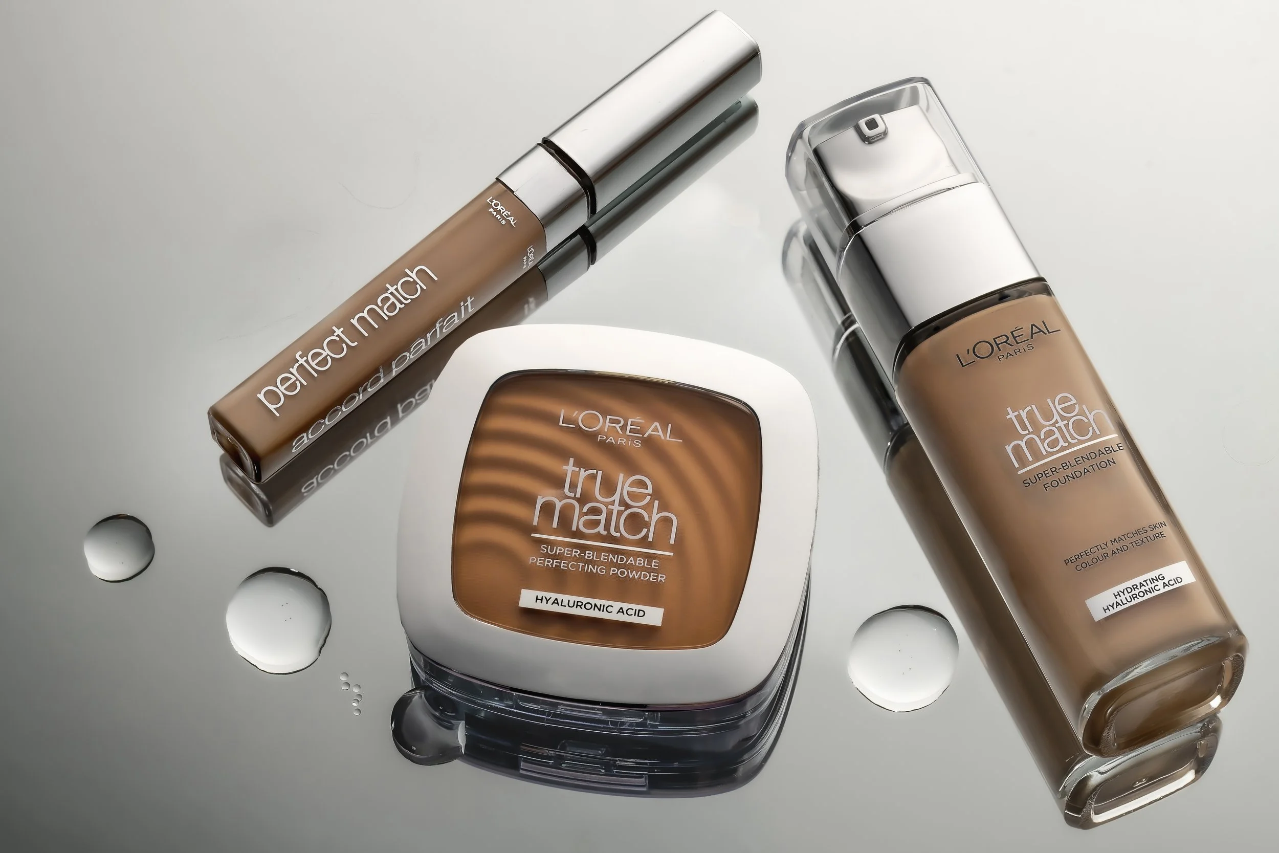 L'Oreal Paris makeup products including True Match foundation, powder, and Perfect Match concealer on a reflective surface with water droplets.