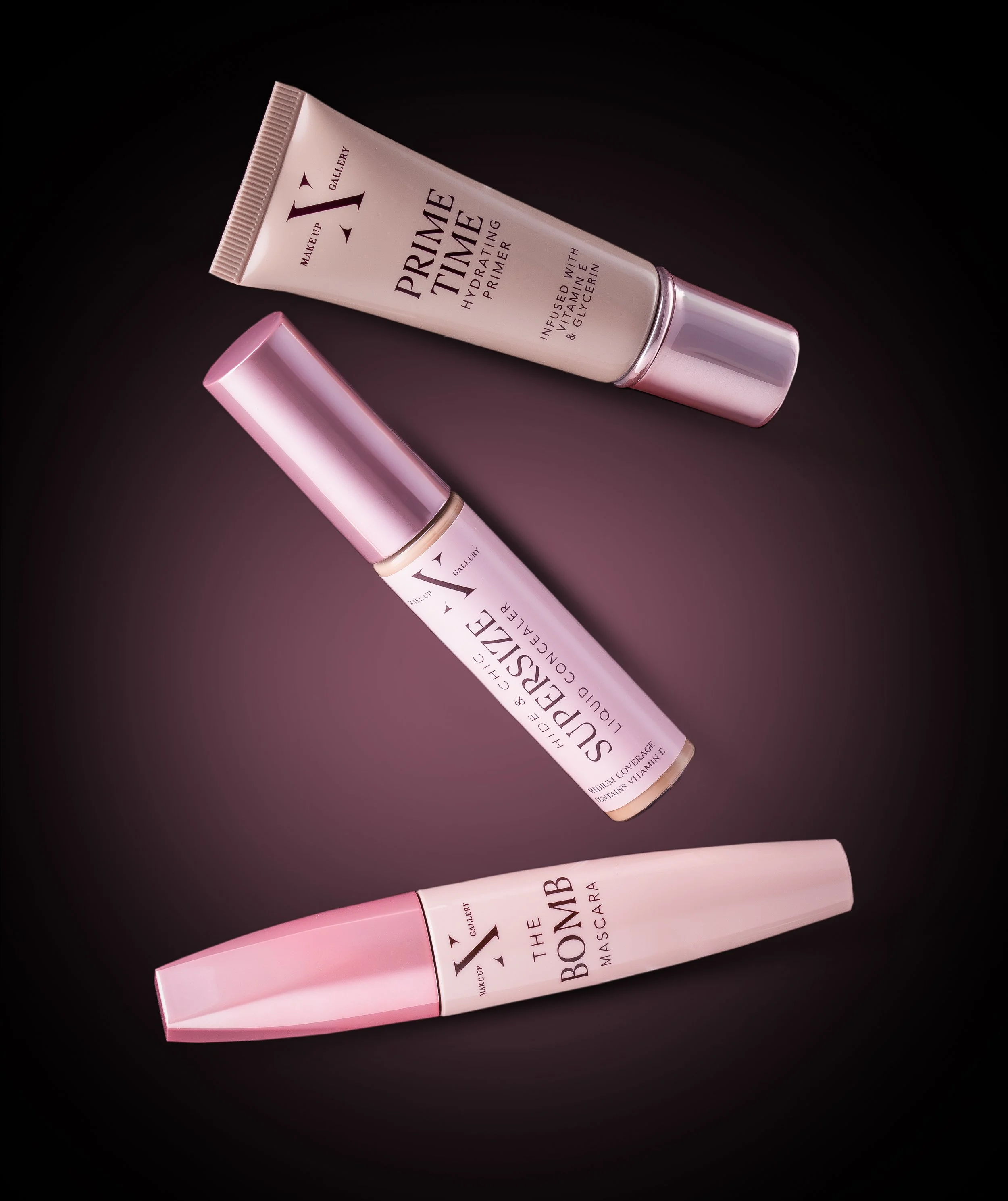 Three makeup products on a dark background, including a primer tube labeled 'Prime Time,' a liquid concealer labeled 'Supersize,' and a mascara tube labeled 'The Bomb.'