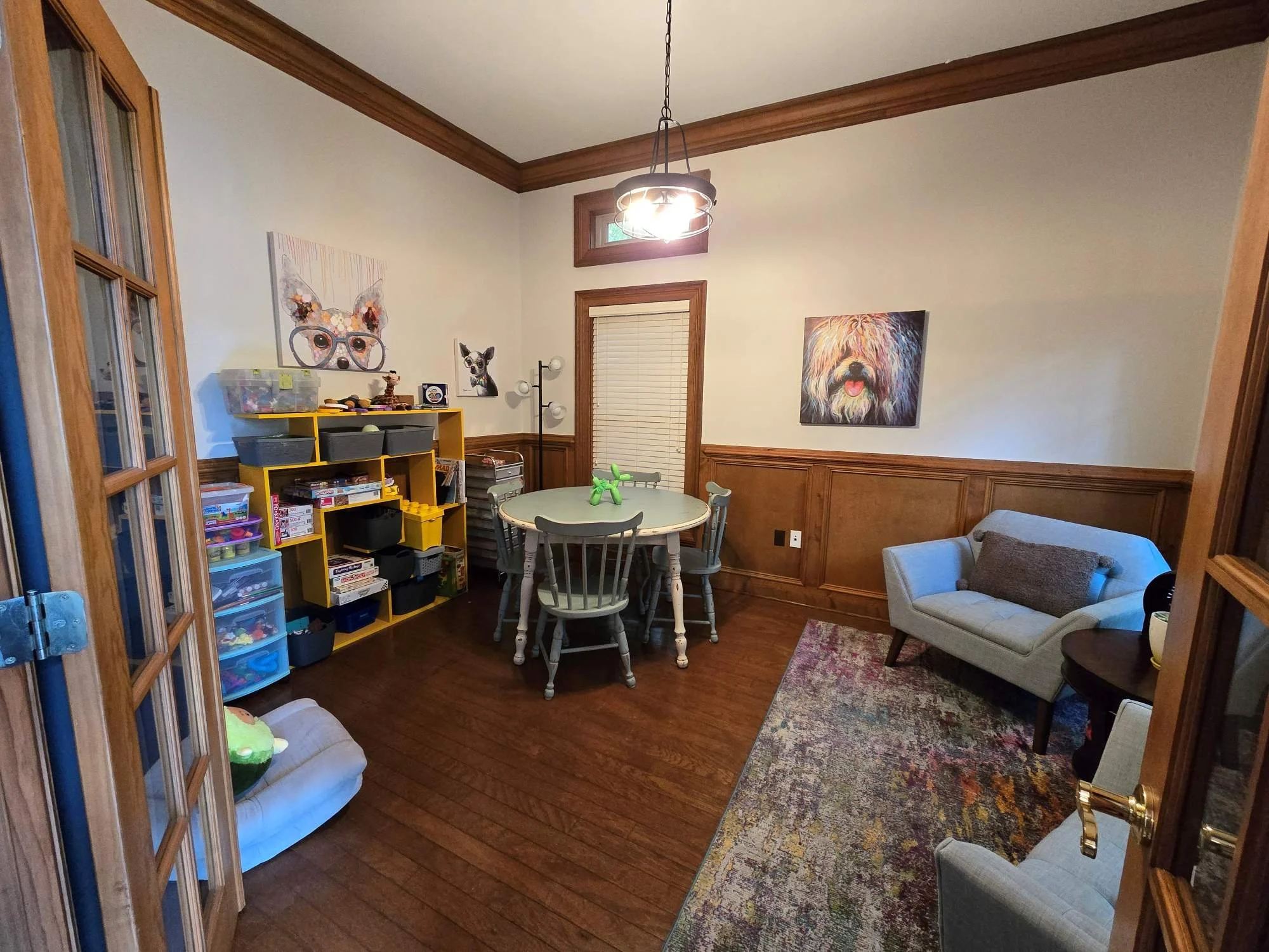 A cozy living room with hardwood floors and wood-paneled walls. It features a white sofa with a gray pillow, a circular dining table with four chairs, a colorful rug, and a bookshelf filled with toys and games. Decor includes animal artwork on the walls and a modern chandelier hanging from the ceiling.