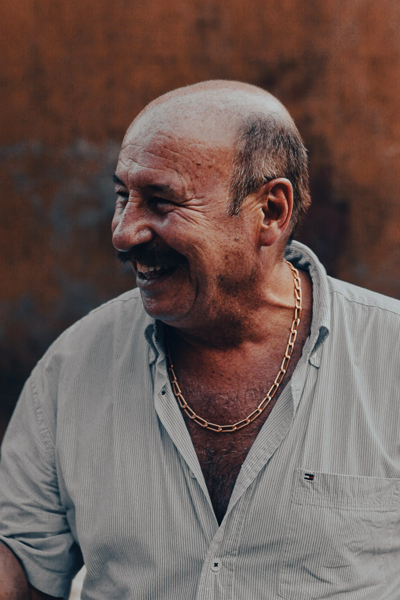 Smiling man wearing a chain necklace and striped shirt against a brown background.