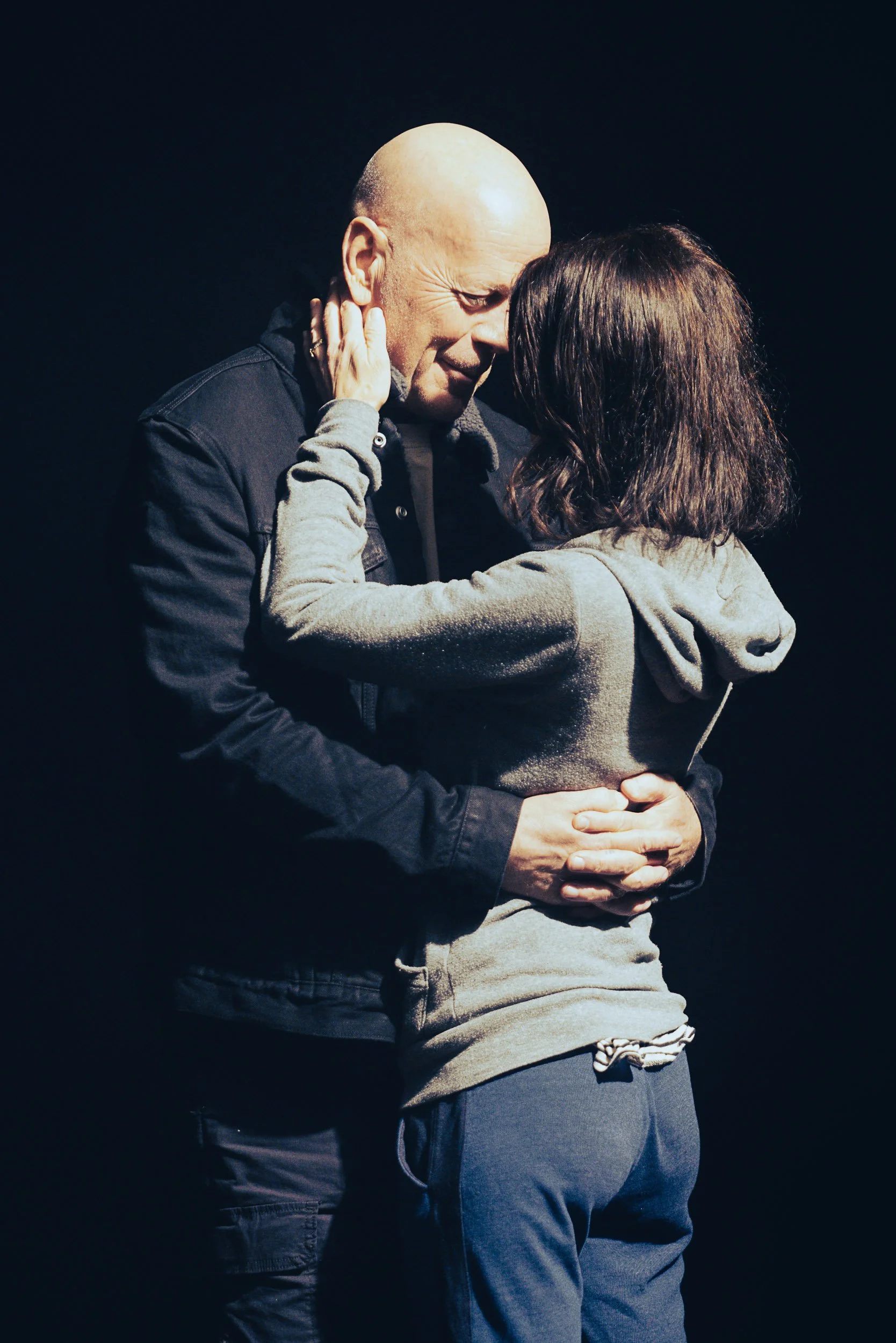 A man and a woman embracing closely in an intimate moment, set against a dark background.