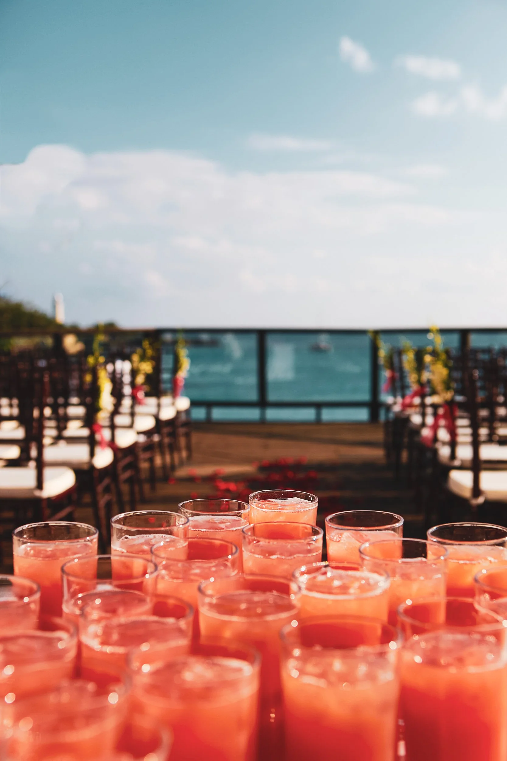 Outdoor wedding setup with rows of chairs and table filled with glasses of pink juice; ocean view in background.