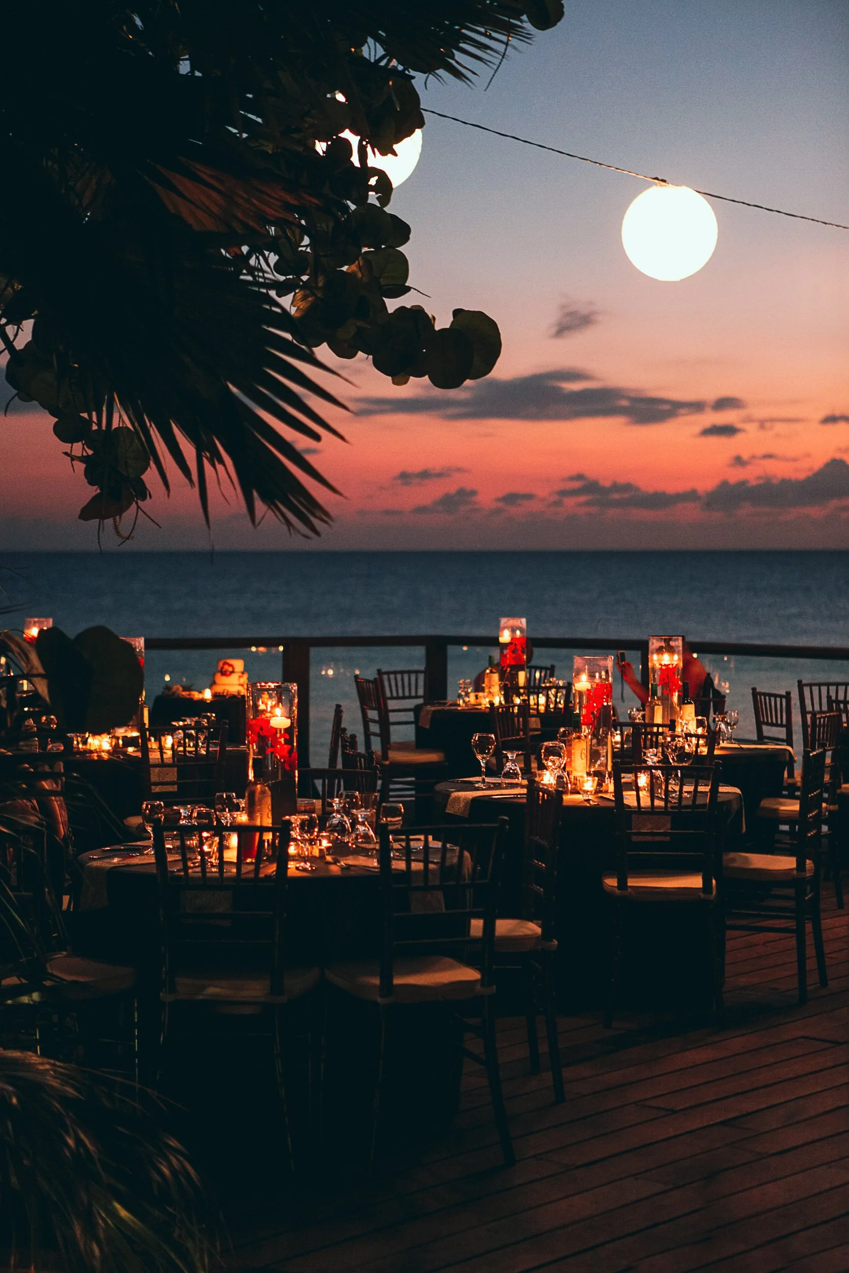 Outdoor seaside dining setup at sunset with round tables, chairs, and candlelit decor, overlooking an ocean view under a pink and purple sky.