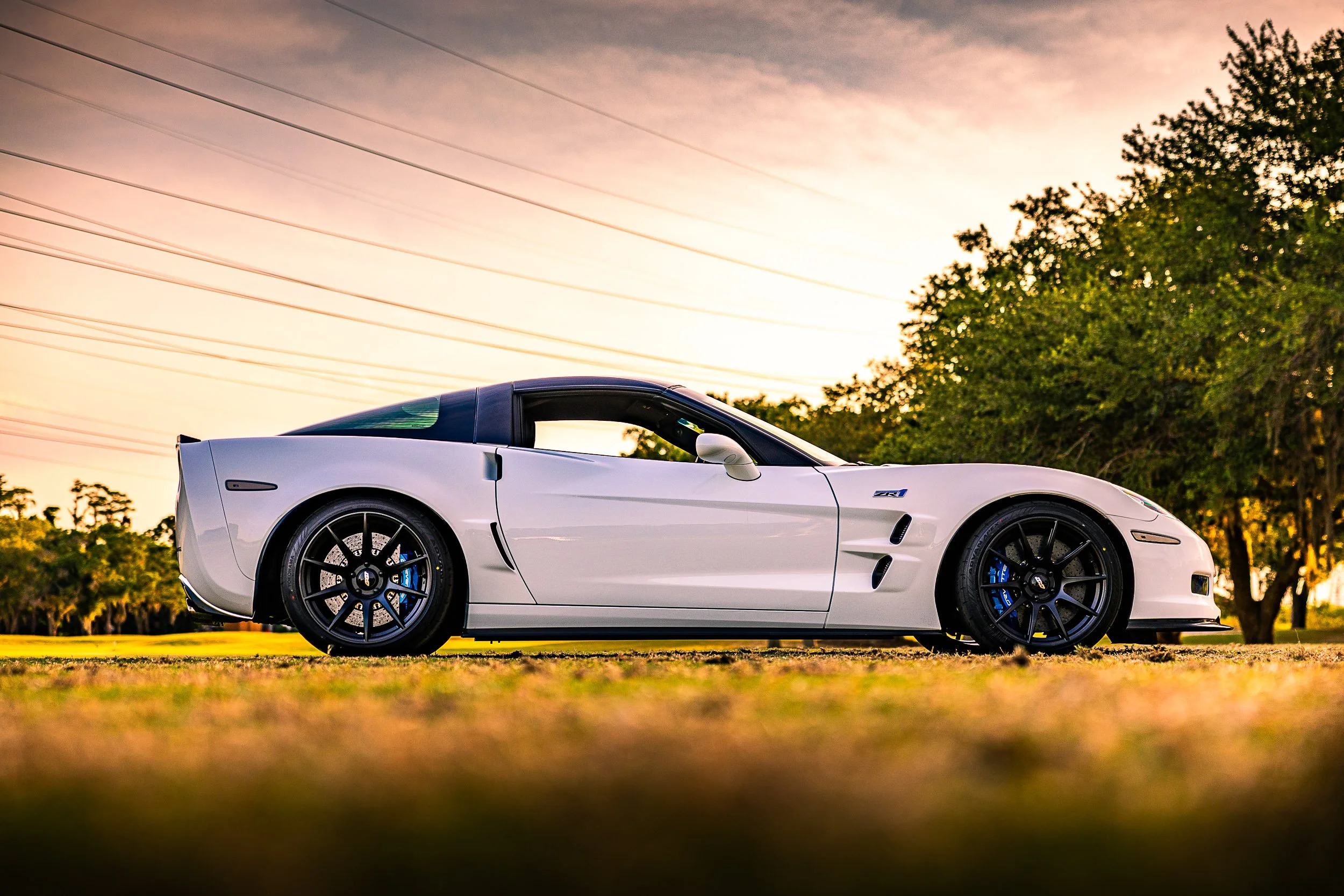 A white sports car parked in a scenic outdoor setting during sunset, with trees and a colorful sky in the background.
