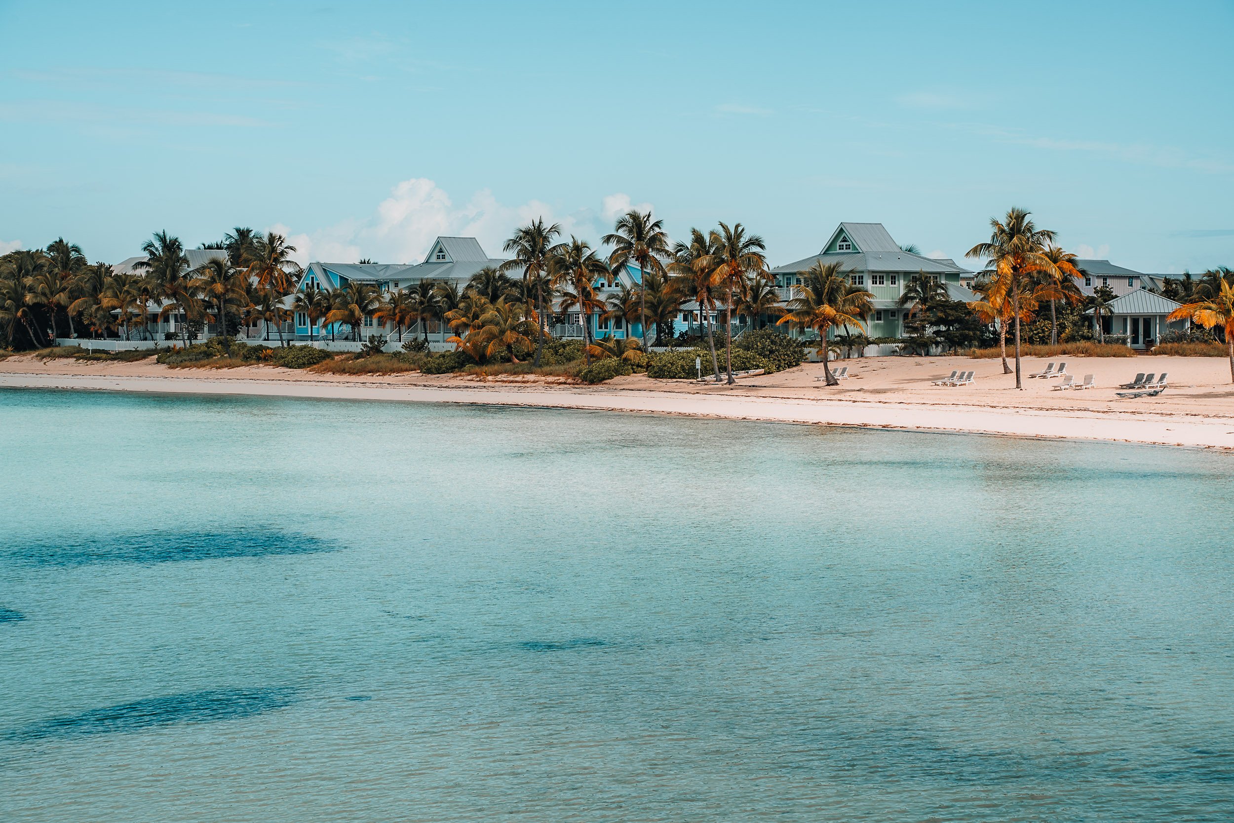 Tropical beach scene with clear turquoise water, sandy shore, and palm trees. Several beach chairs and resort buildings in the background under a blue sky.