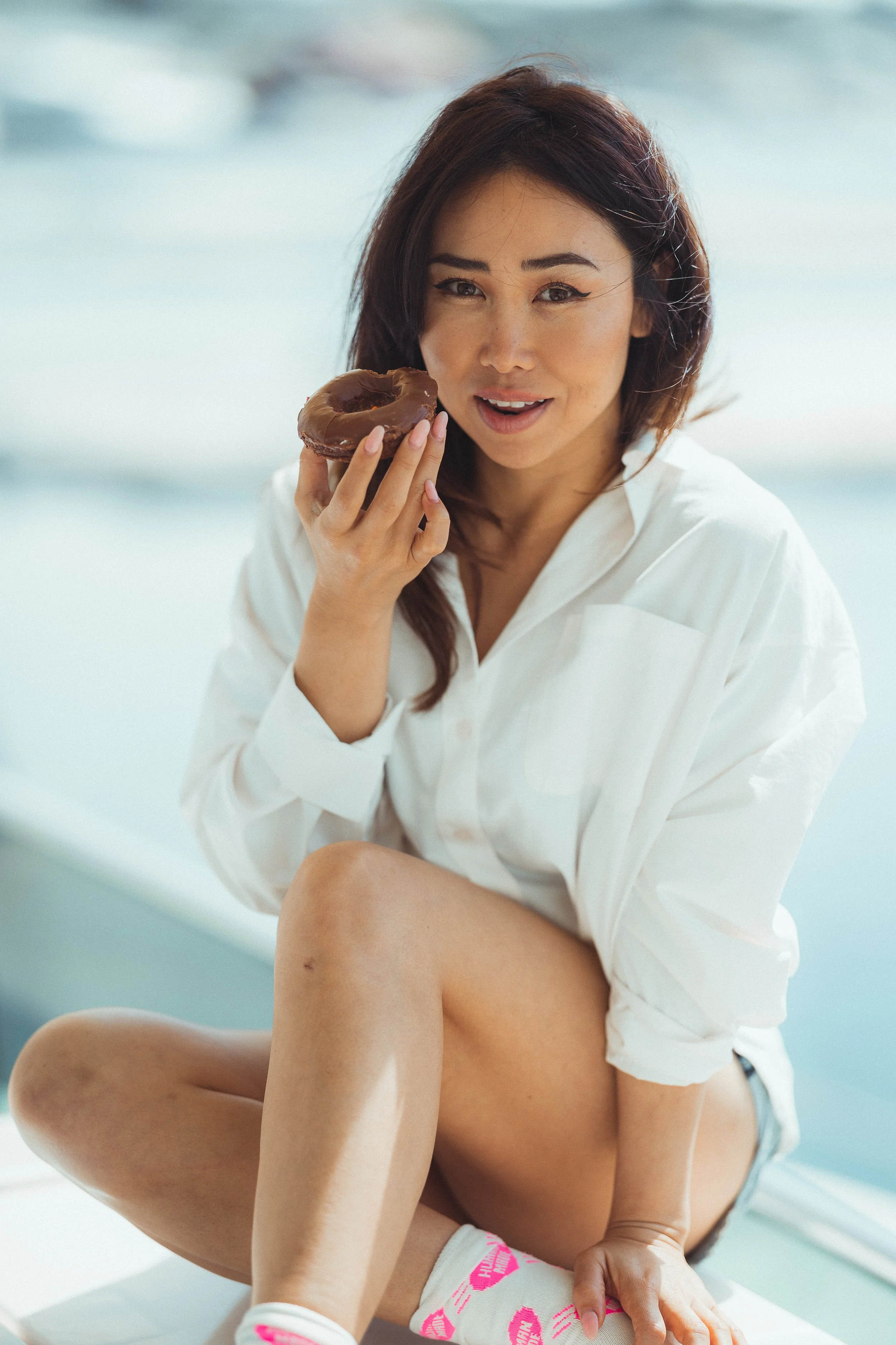 A woman with dark hair and light to medium skin tone, wearing a white shirt, holding a chocolate glazed donut, smiling.