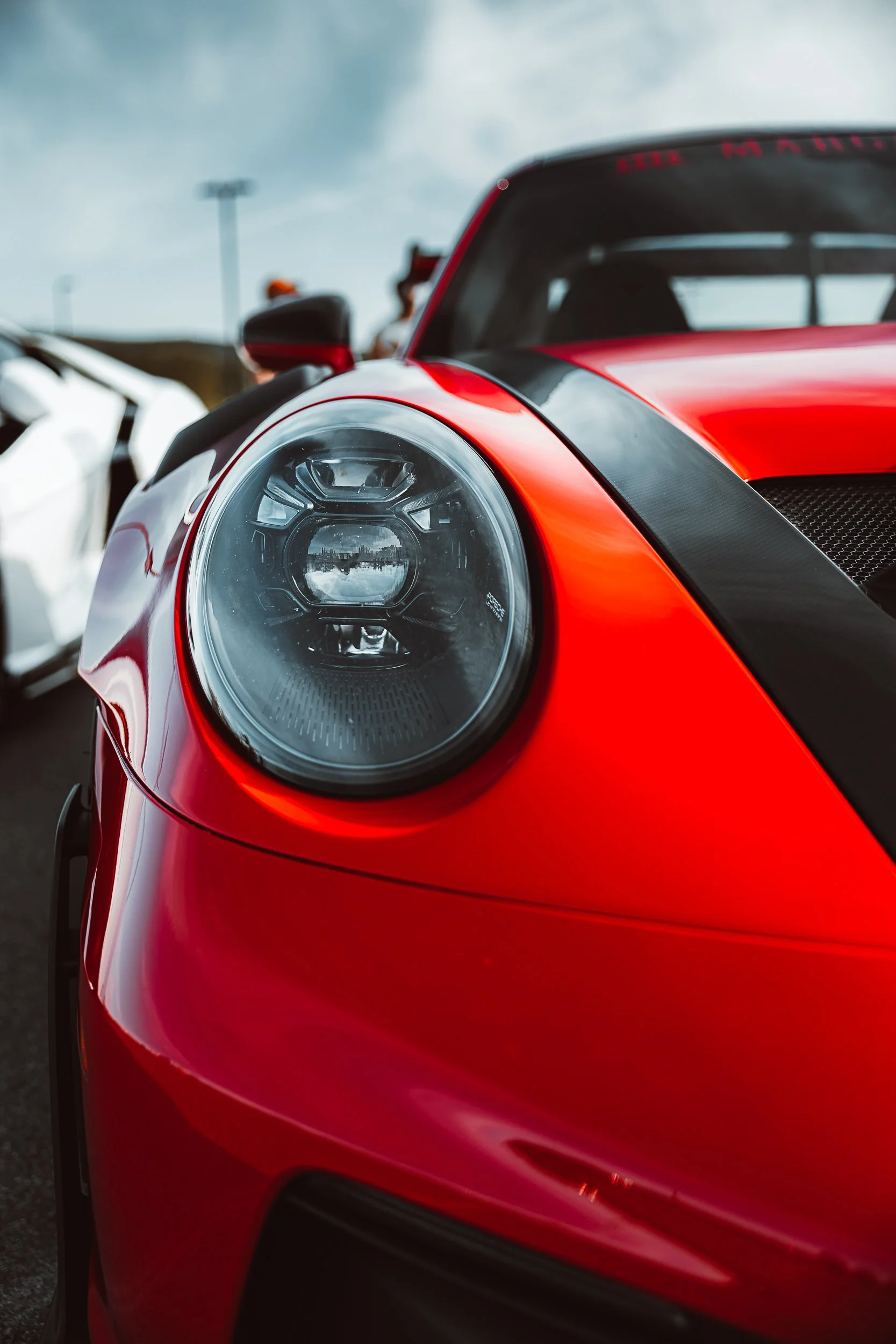 Close-up of the front part of a red sports car, focusing on the headlight and bumper, with a blurred background of cloudy sky and people.
