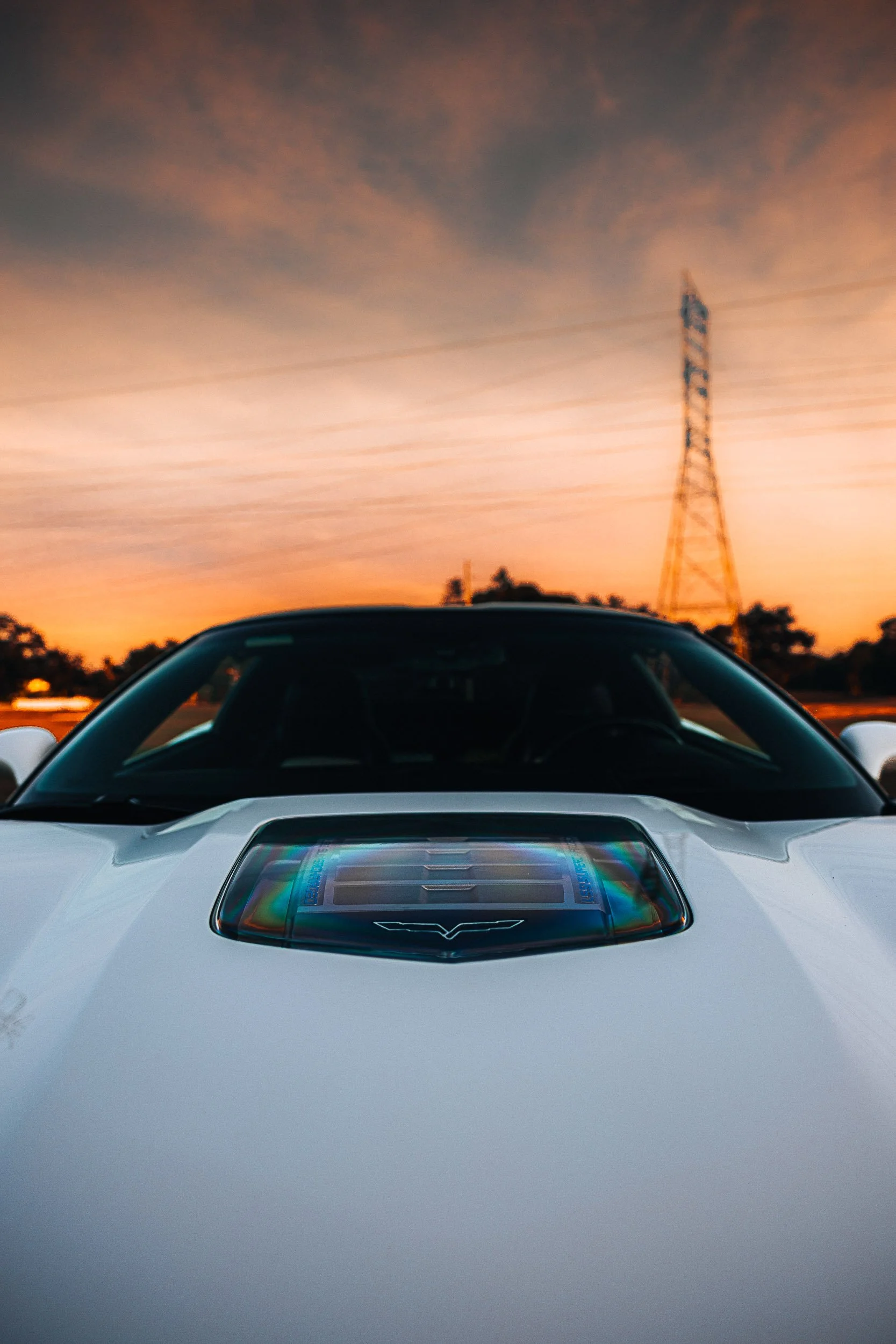 Close-up of a white sports car's hood with a transparent engine cover, under a dramatic evening sky with a telecommunications tower in the background.
