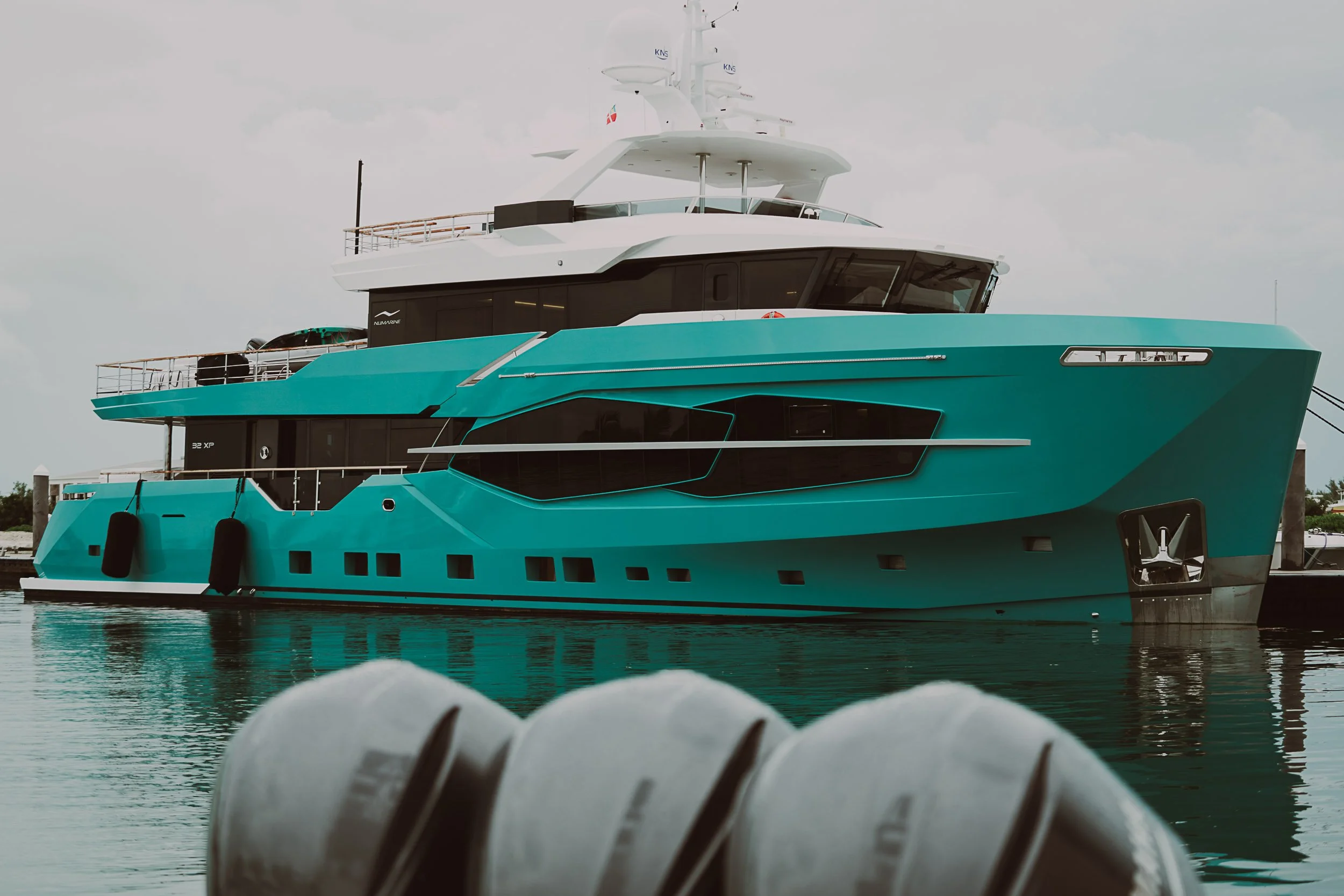 A large, teal-colored yacht docked in a marina, featuring a modern design and multiple decks, with inflatable boats in the foreground.