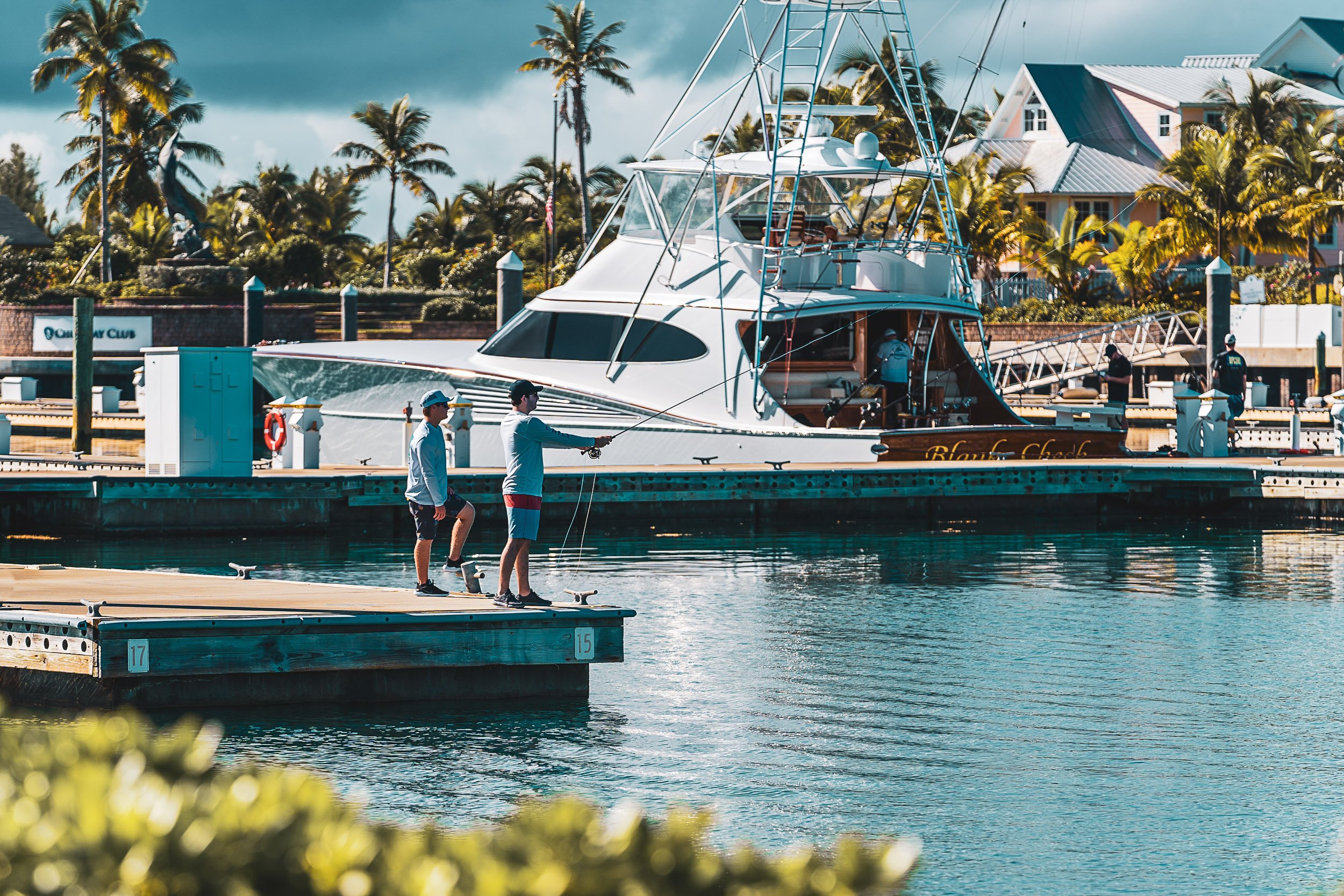 Two people fishing on a dock near a yacht, surrounded by palm trees and water, with a blue sky overhead.