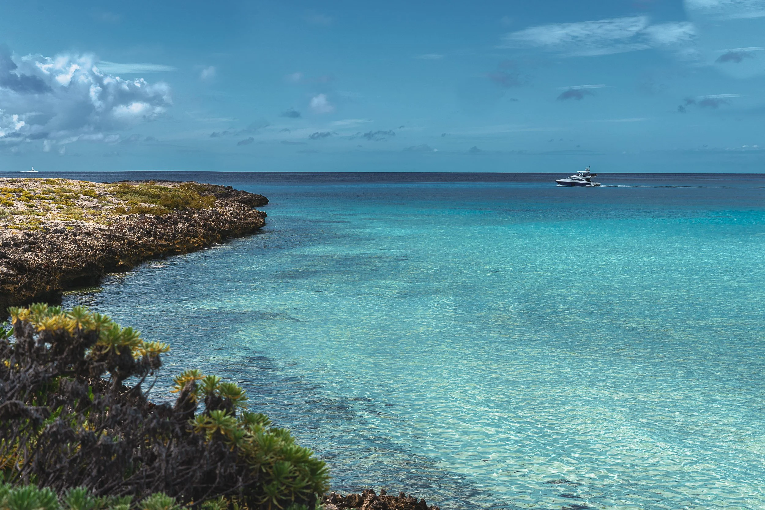 Clear turquoise sea with a rocky shoreline and vegetation in the foreground, a boat on the water, and a blue sky with clouds.