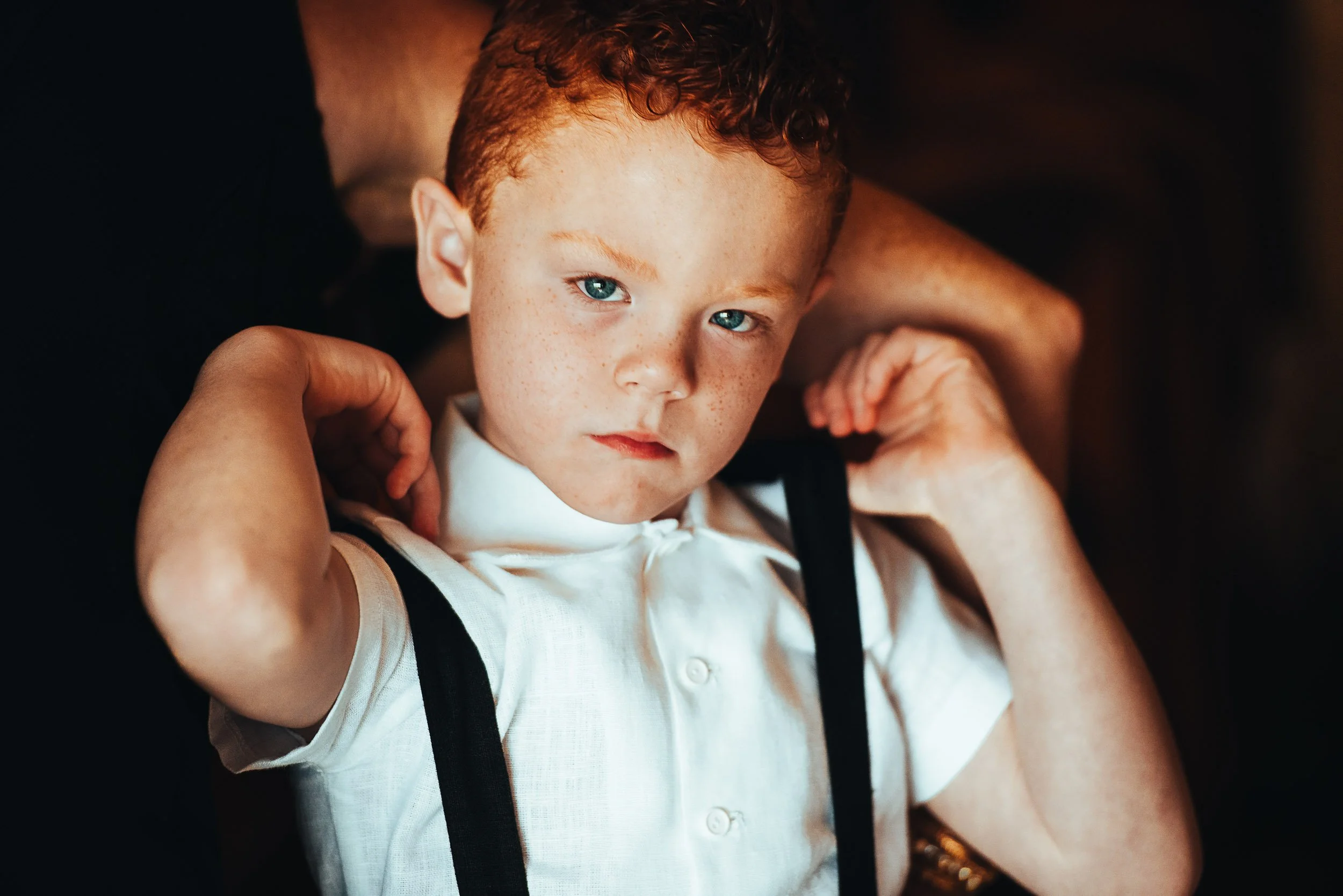 Young child with red hair, wearing a white shirt and suspenders, looking serious.