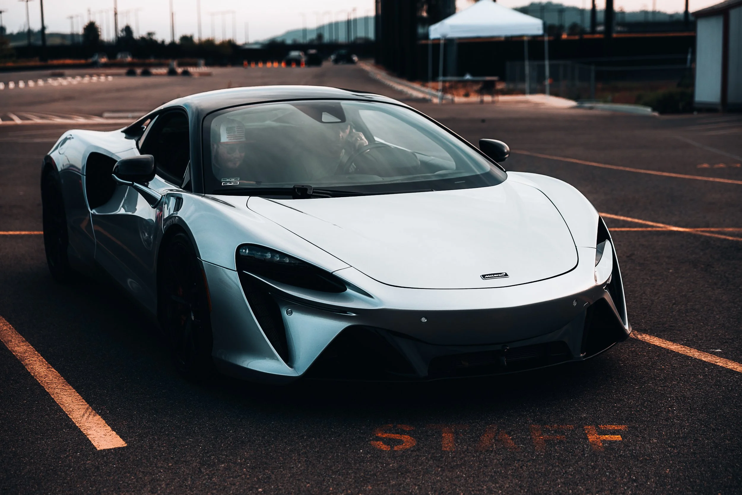 A silver McLaren sports car parked in an empty parking lot at dusk, with two people inside, one wearing sunglasses and the other holding a can, both facing forward.