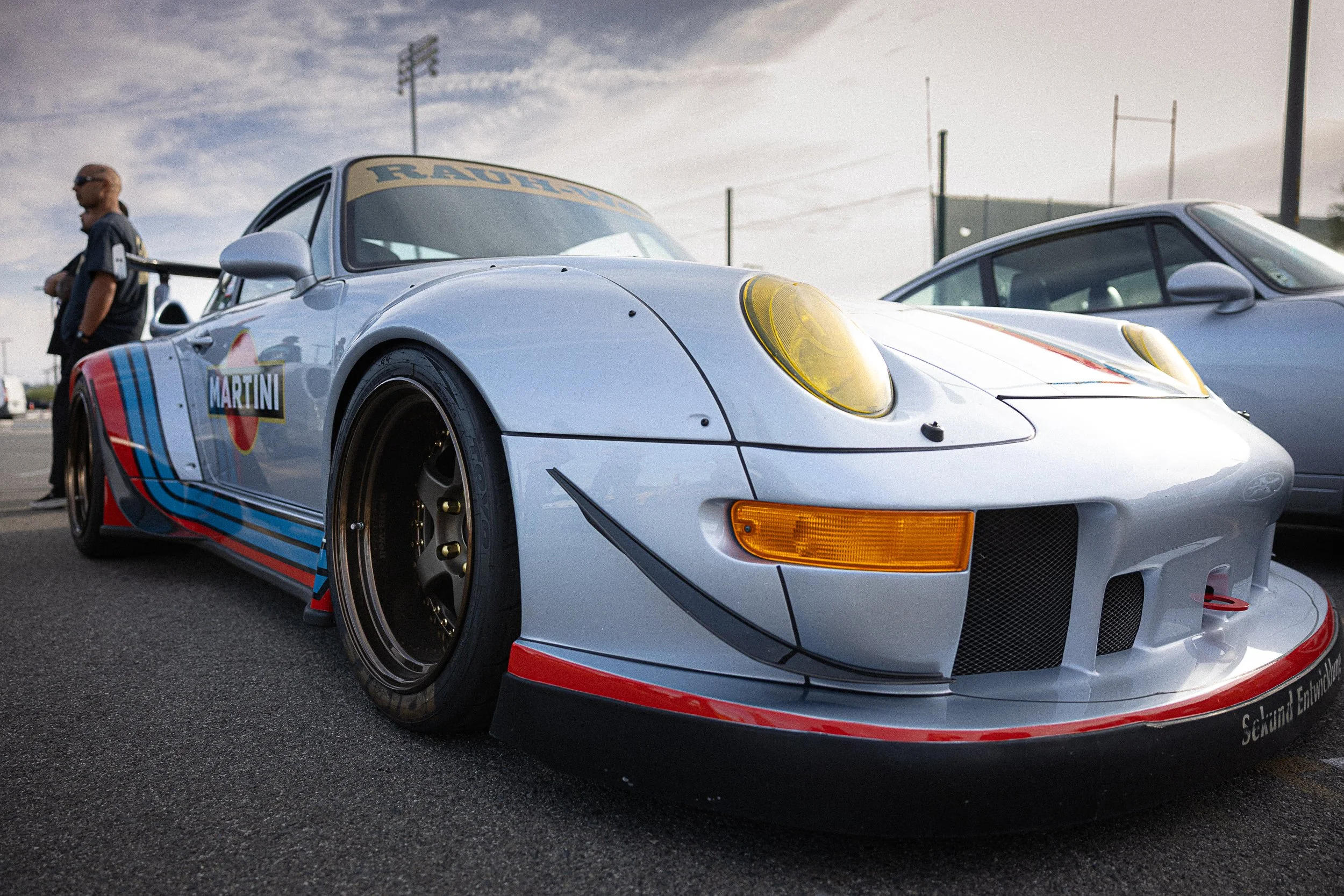 A vintage race car with Martini racing livery parked at a race track with a person walking in the background.