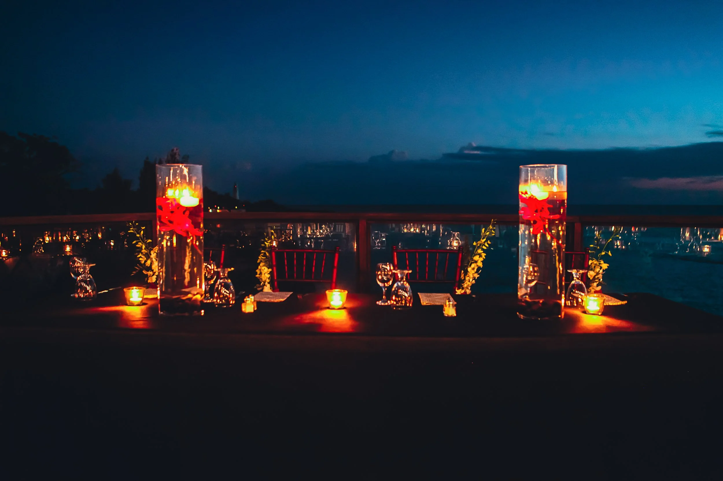 Romantic candlelit dinner table with tall glass vases and glowing candles against a dark night sky backdrop.