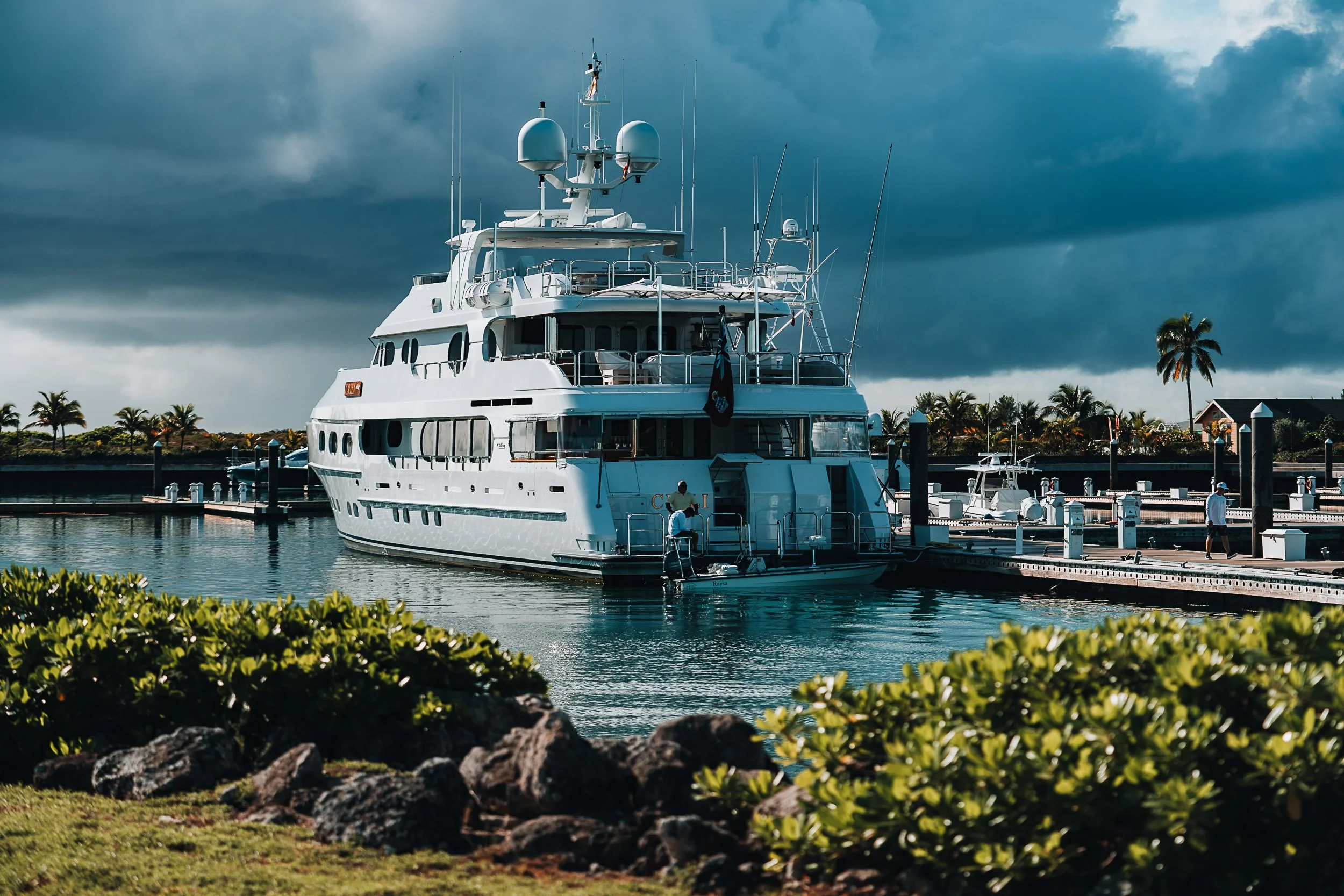 Luxury yacht docked at a marina with cloudy sky and palm trees in the background.