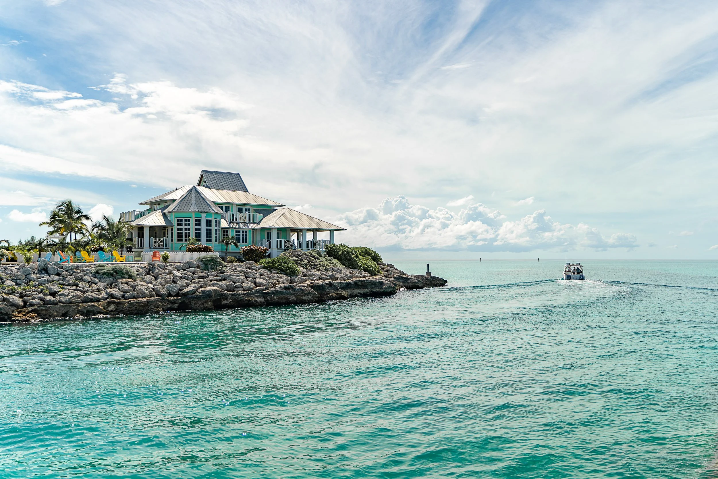 Beachfront house with Adirondack chairs, palm trees, and a boat on turquoise water under a cloudy sky.