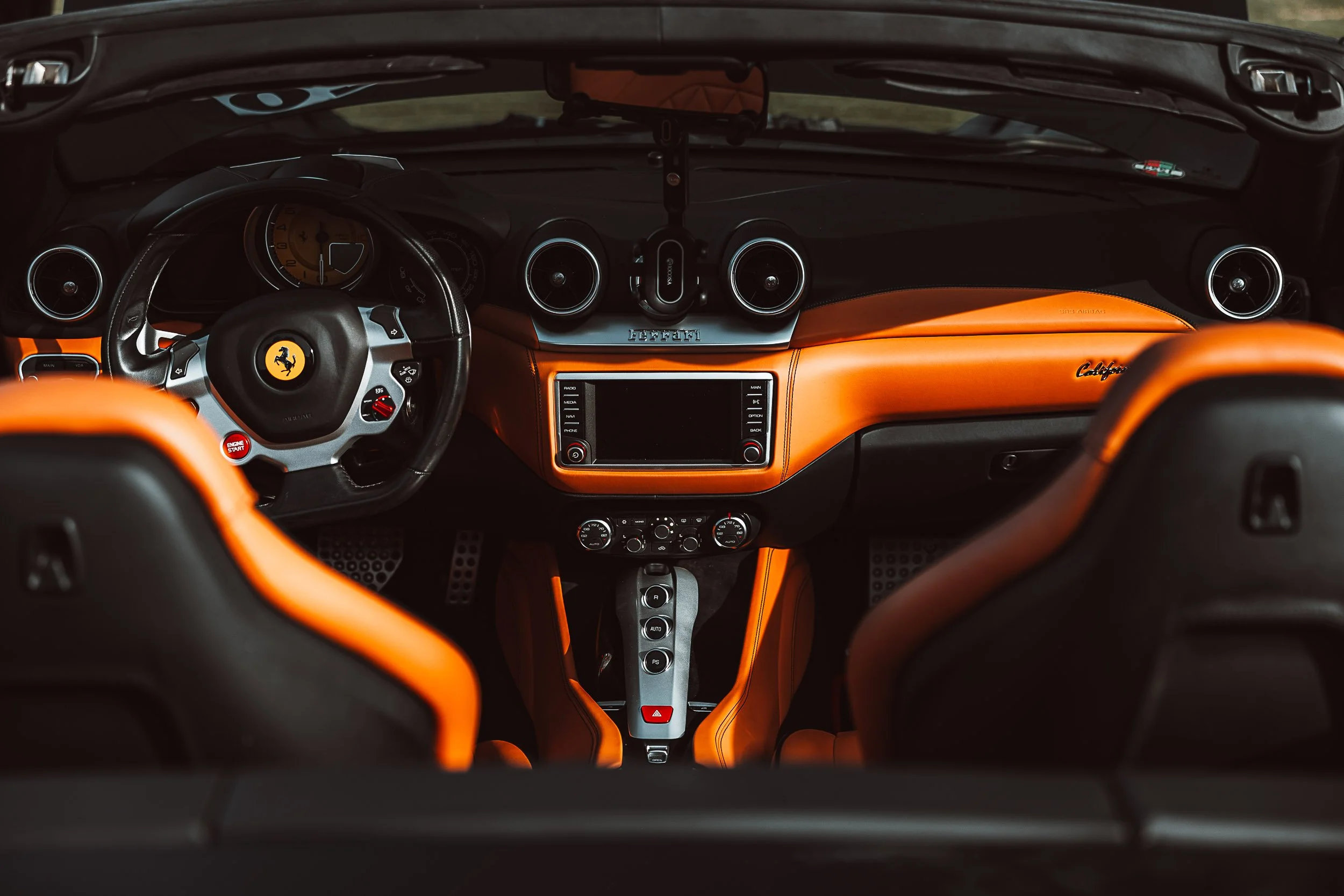 Interior of a luxury sports car with orange and black leather, featuring a steering wheel with a yellow emblem, central console with controls and a display."