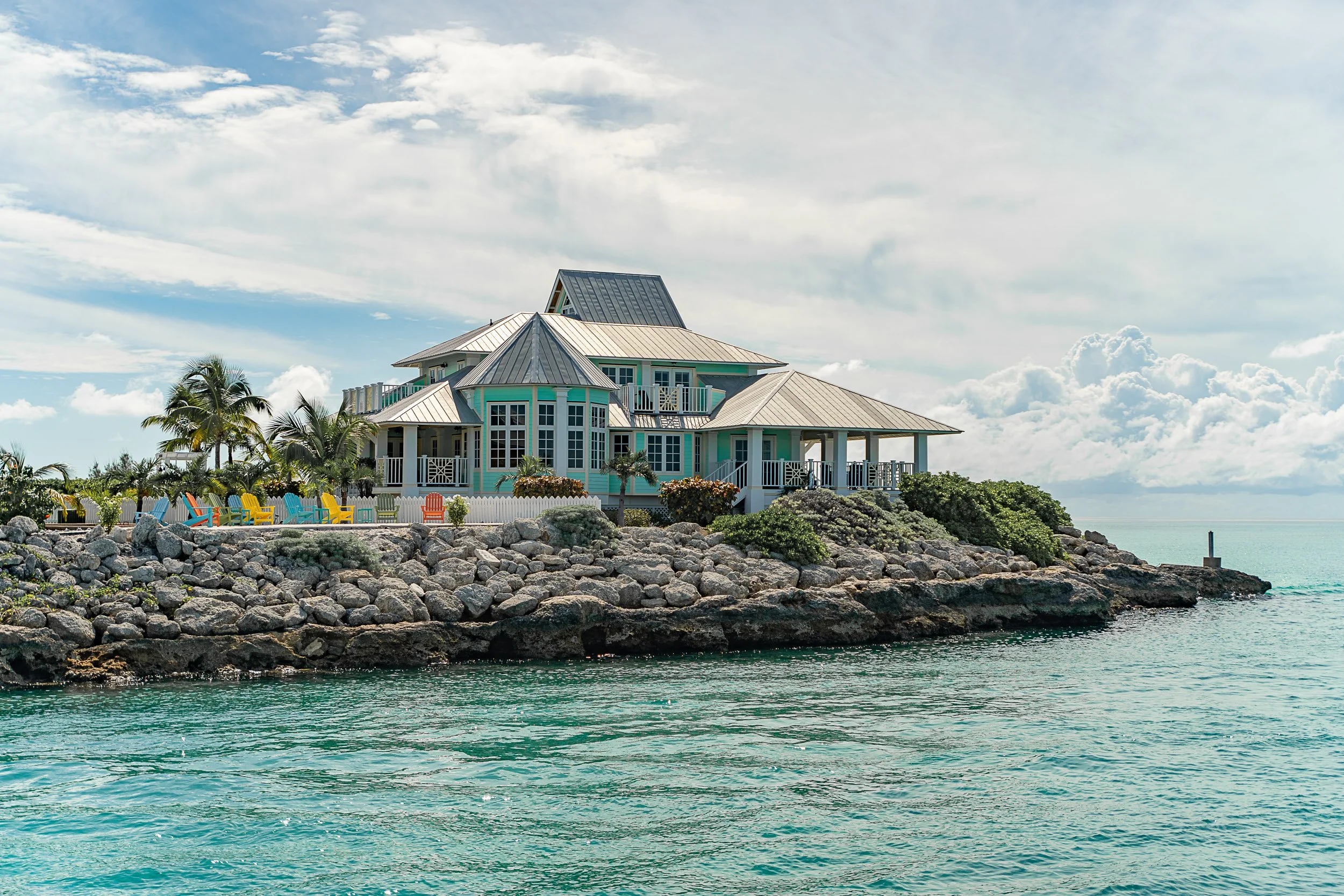 Beachside house with teal accents, Adirondack chairs, and palm trees on rocky shoreline