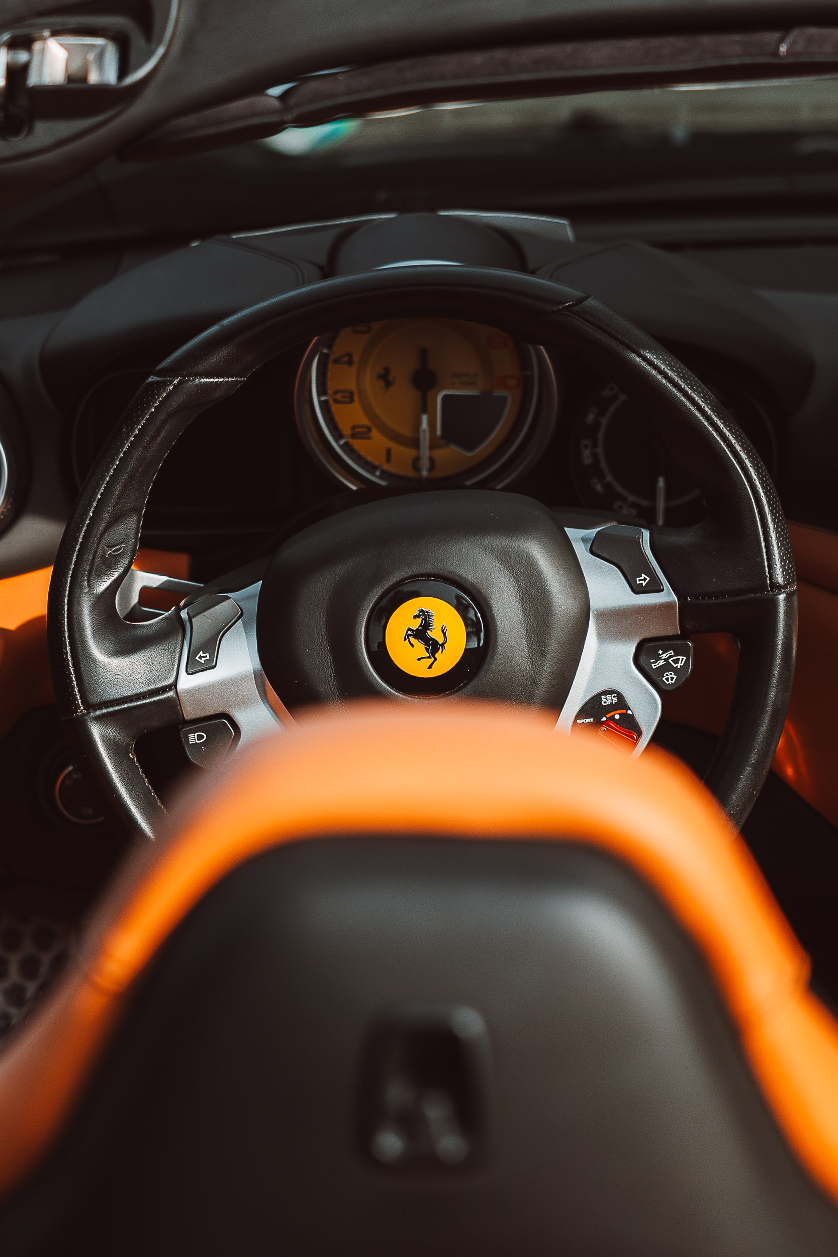 Interior view of a car with a black steering wheel featuring a yellow prancing horse logo and orange accents, with a yellow speedometer in the background.