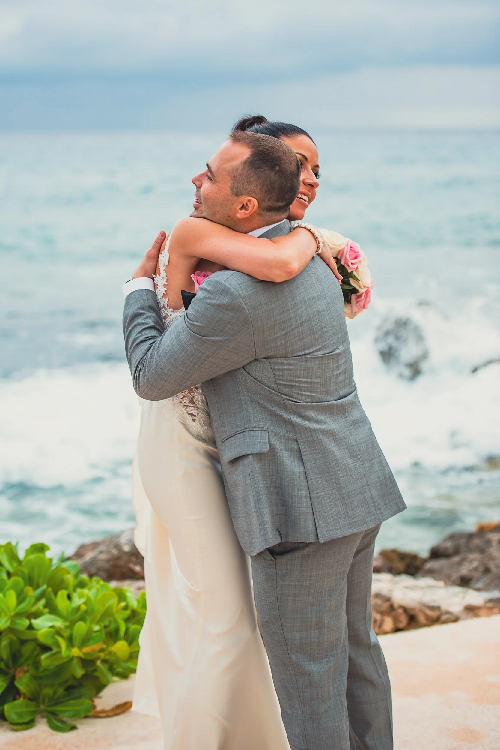 A couple in formal attire hugging by the ocean, with pink roses visible.