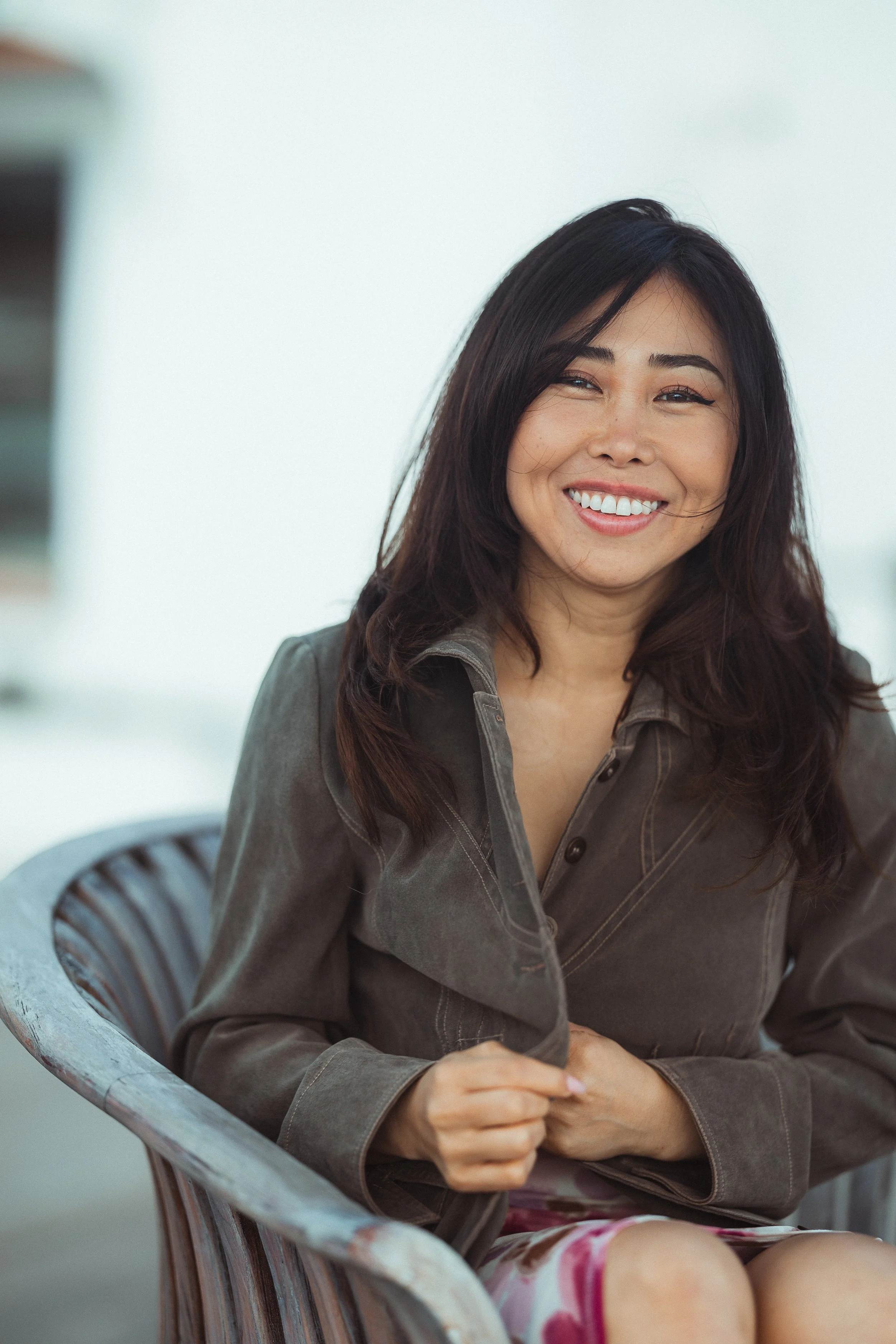A woman sitting in a weathered plastic chair, smiling and looking at the camera, with wind-blown hair, wearing a gray jacket and colorful shorts, with a cloudy sky in the background.
