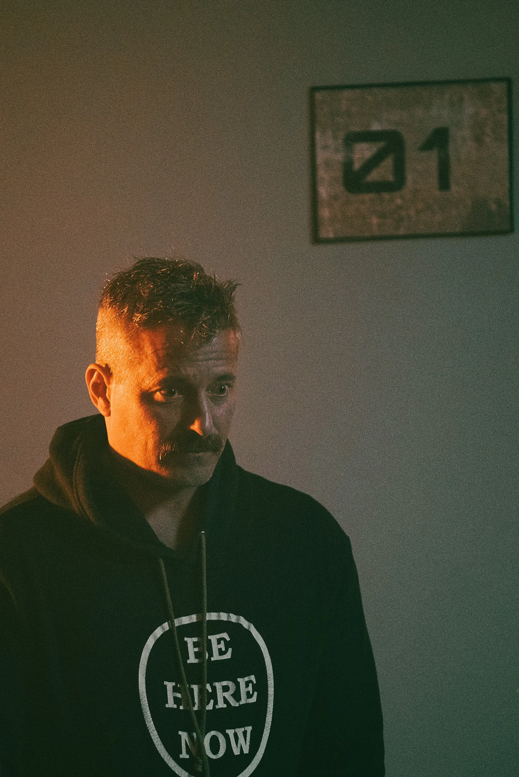Man with mustache in dimly lit room wearing "Be Here Now" hoodie standing in front of a wall with a sign displaying "01."