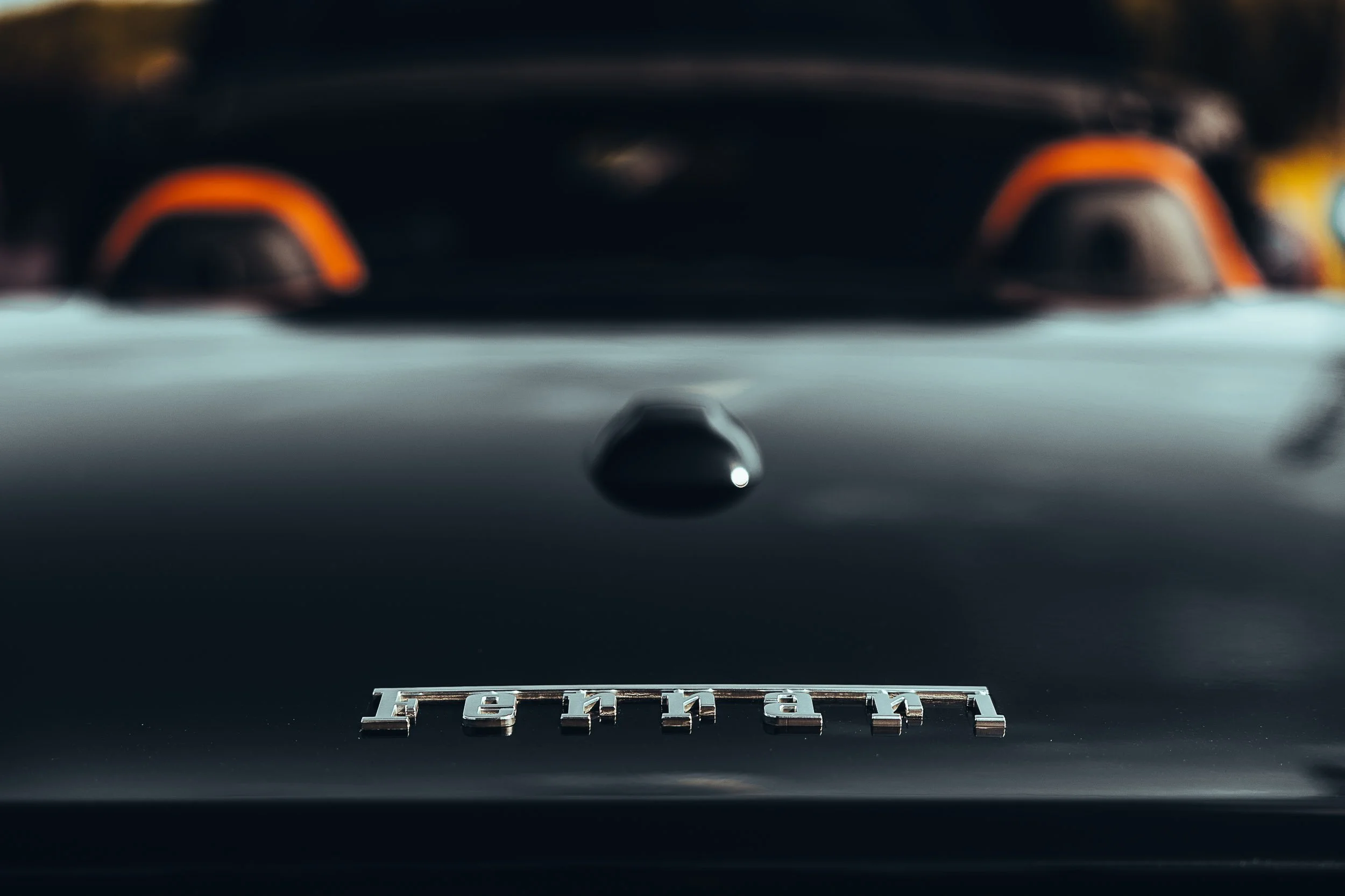 Close-up of a Ferrari logo on a dark car hood with blurred background, featuring orange accents.