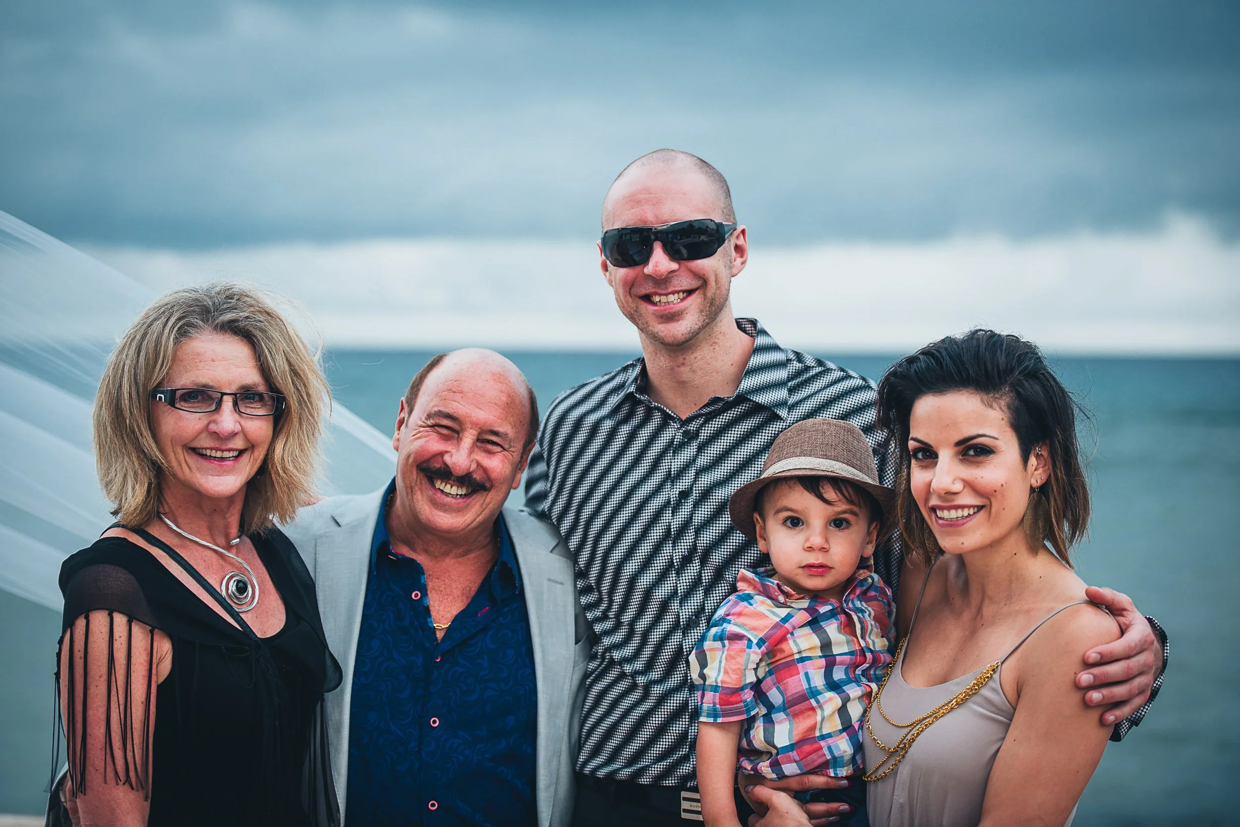 Group of five people smiling at the camera, including a man in sunglasses, a woman holding a child wearing a hat, and two adults with glasses, with a cloudy ocean backdrop.