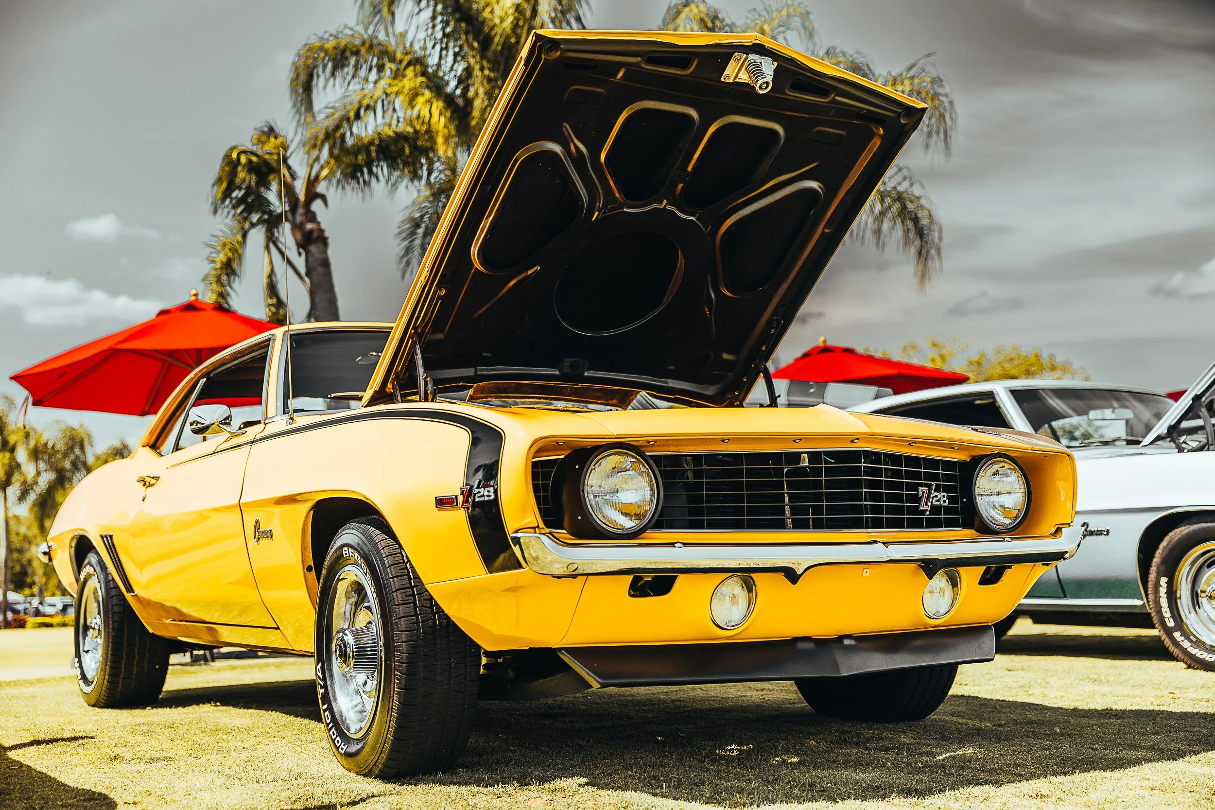 Yellow classic muscle car with hood open at a car show, palm trees and red umbrellas in background.