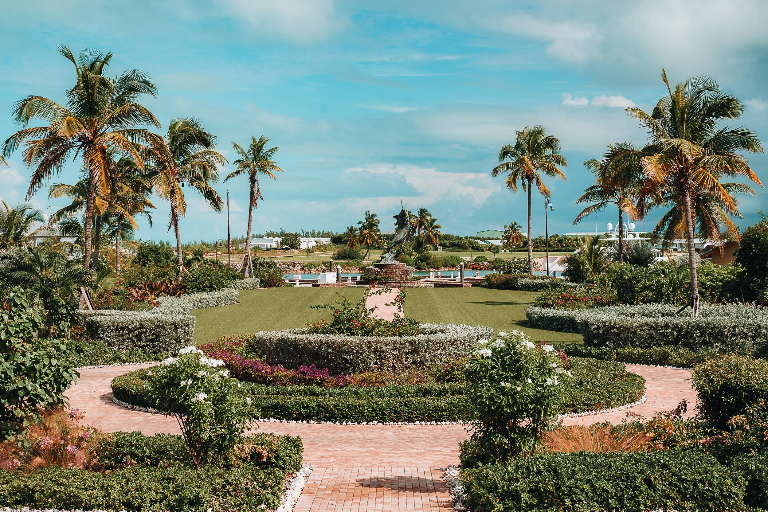 Tropical garden with palm trees, lush greenery, and a central pathway leading to a fountain under a blue sky.