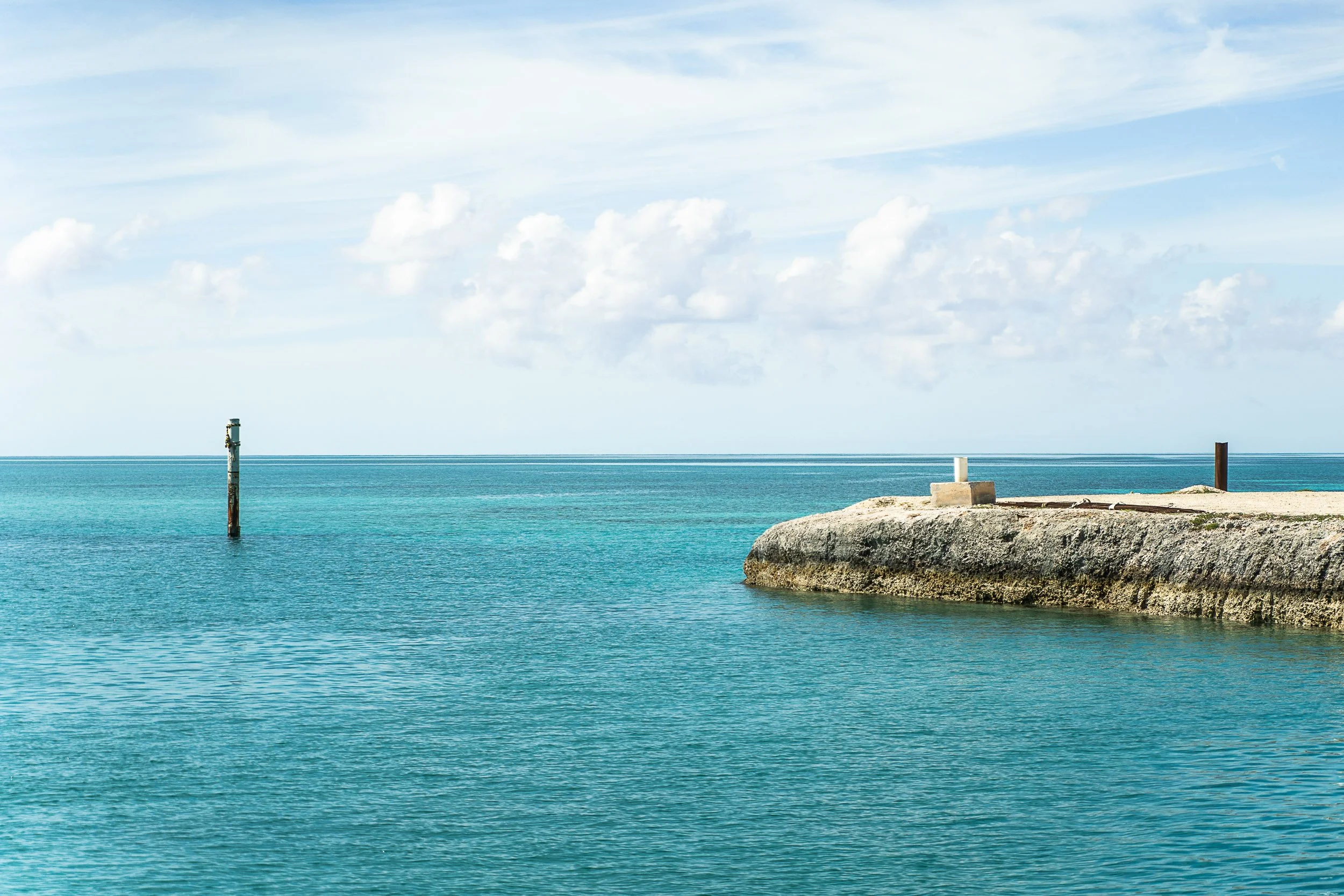 Clear blue ocean with a small rocky pier extending into the water; a single post or marker is visible in the distance; fluffy clouds in the sky.