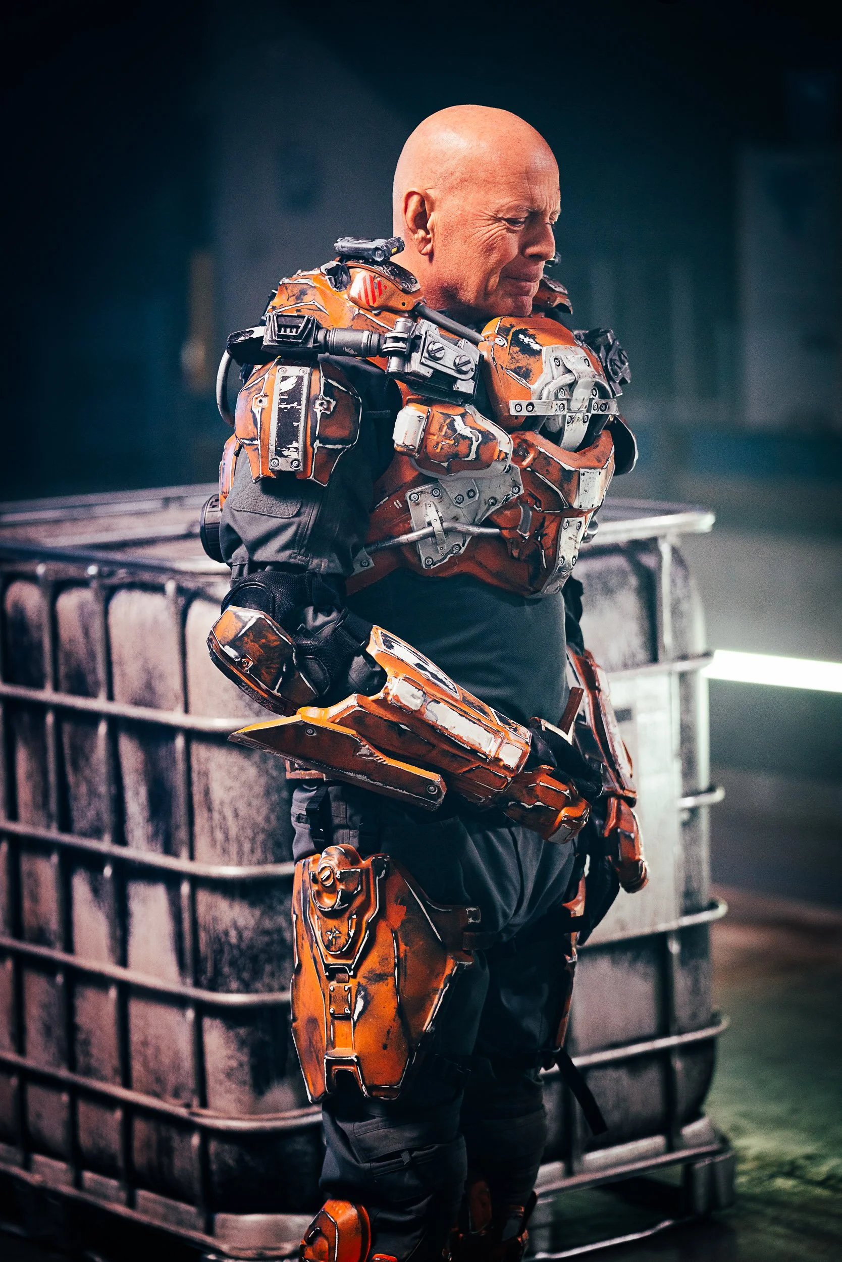 A bald man wearing a futuristic orange armored suit stands next to a large metal crate.