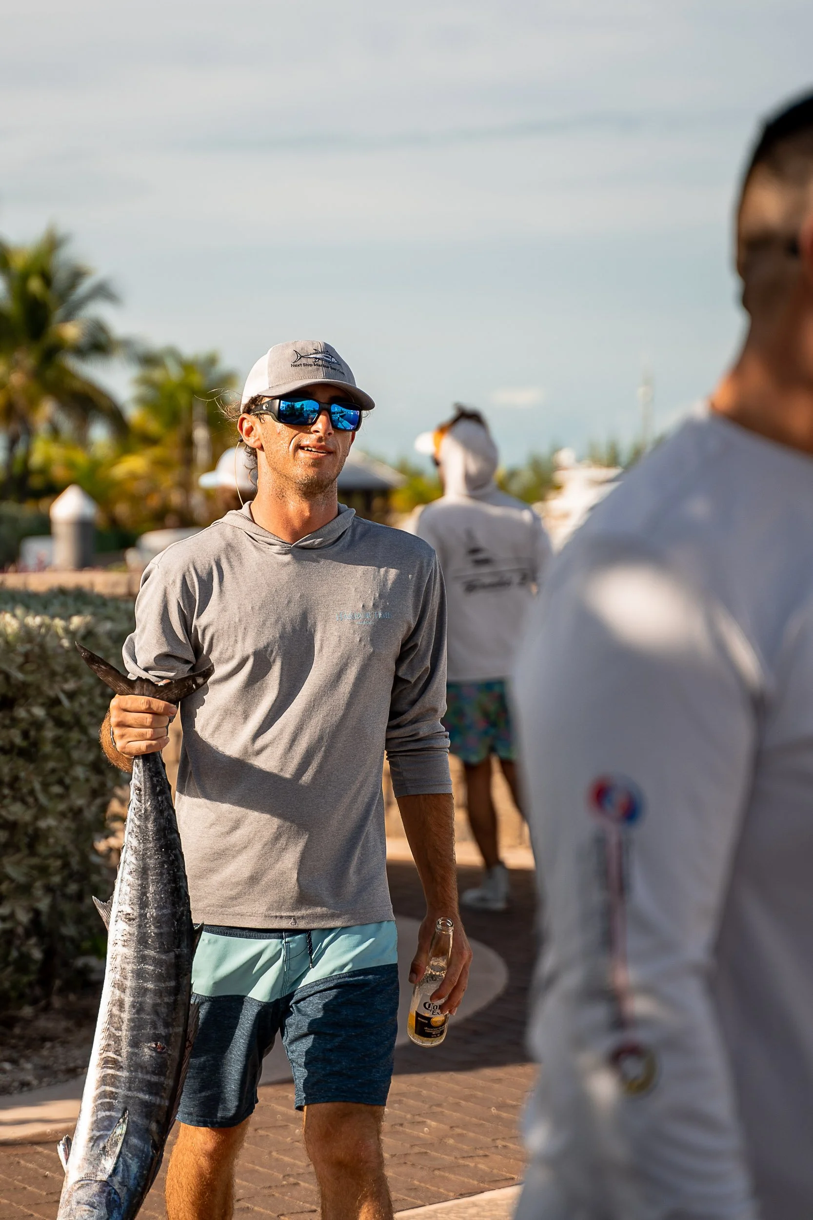 Man holding a fish and a beverage outdoors in sunny weather, wearing sunglasses and a cap, with palm trees in the background.