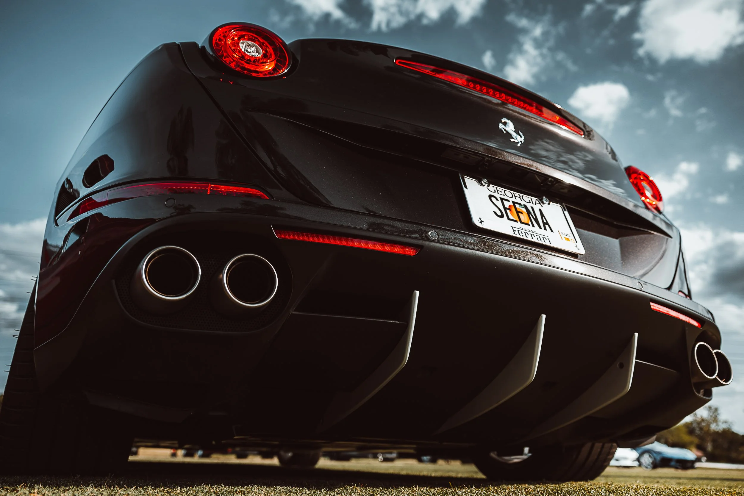 Low-angle view of the rear of a black Ferrari with dual exhaust pipes and a personalized Georgia license plate, against a cloudy sky backdrop.